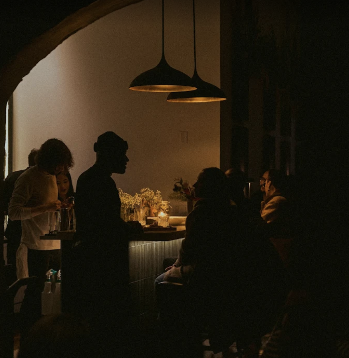 Dimly lit restaurant interior with silhouetted people sitting and talking near a bar, warm hanging lamps, and soft ambient lighting.