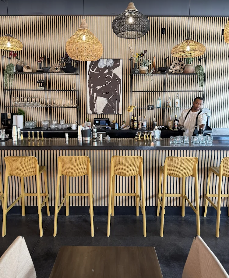 A modern bar with a black countertop and five yellow bar stools. Behind the bar, a bartender stands near shelves with bottles and decor. The wall features wooden slats, decorative lamps, framed artwork, and hanging plants.