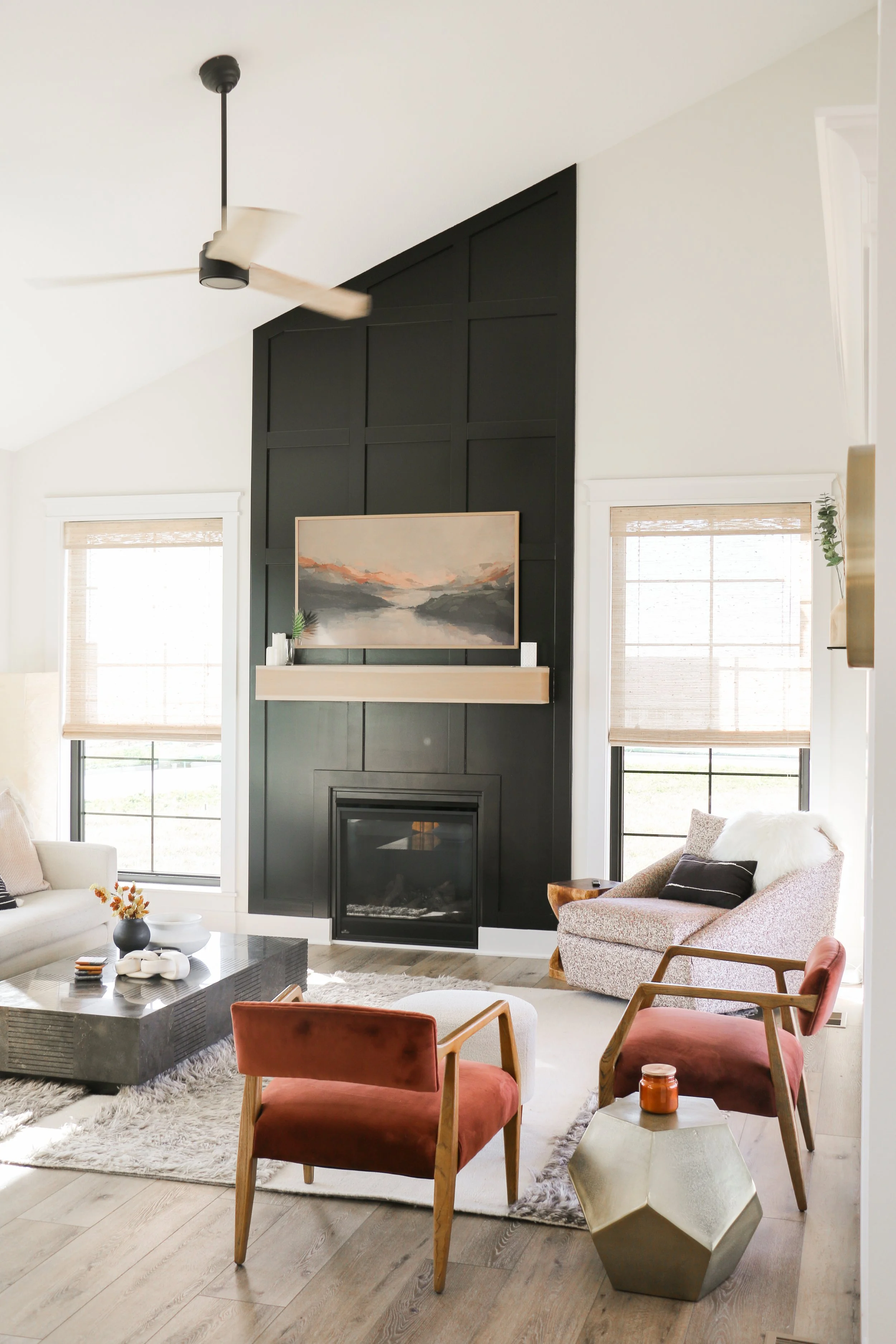 Modern living room with high ceiling, featuring a dark accent wall and fireplace. Decor includes a landscape painting, wooden furniture, and a geometric coffee table. Natural light filters through large windows with light-colored blinds. A ceiling fan and cozy seating complete the room.