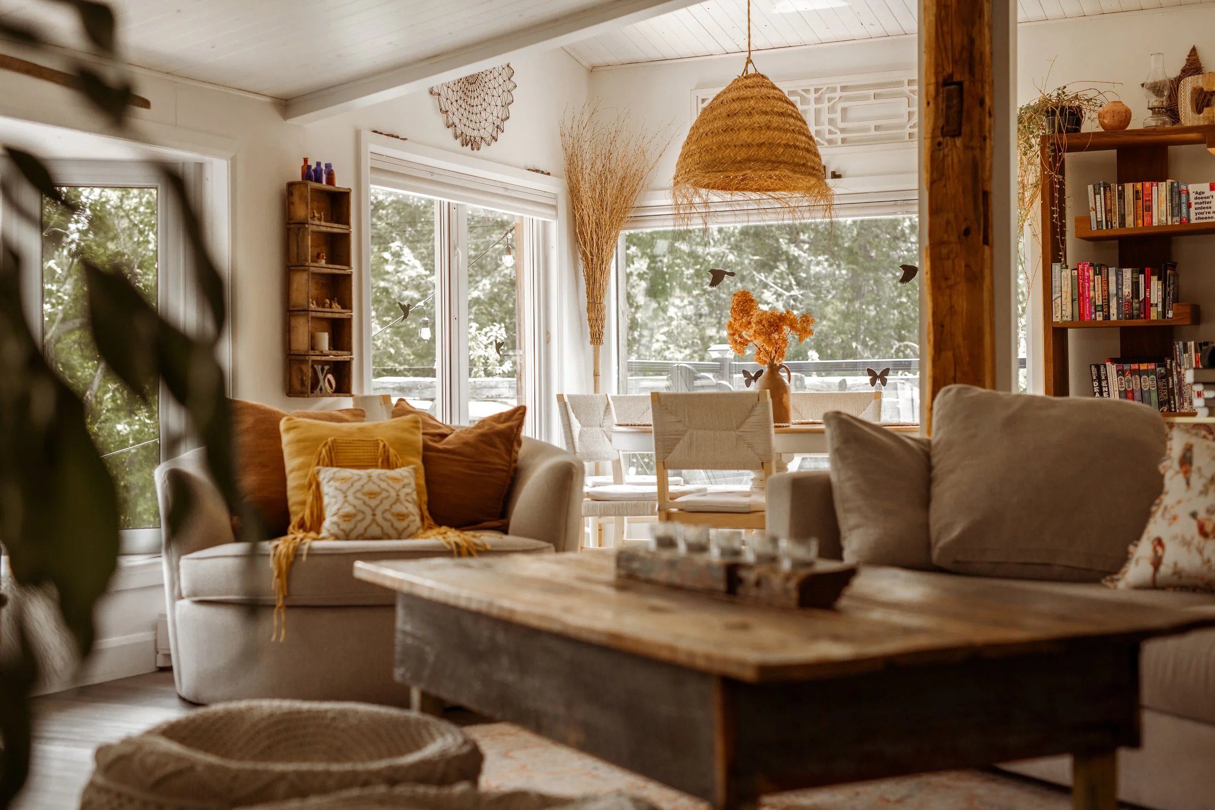 Cozy living room with beige sofa, mustard and brown pillows, and a rustic wooden coffee table. Large windows with greenery outside, a dining area with a vase of orange flowers, decorative wall hangings, a woven pendant light, and a bookshelf filled w