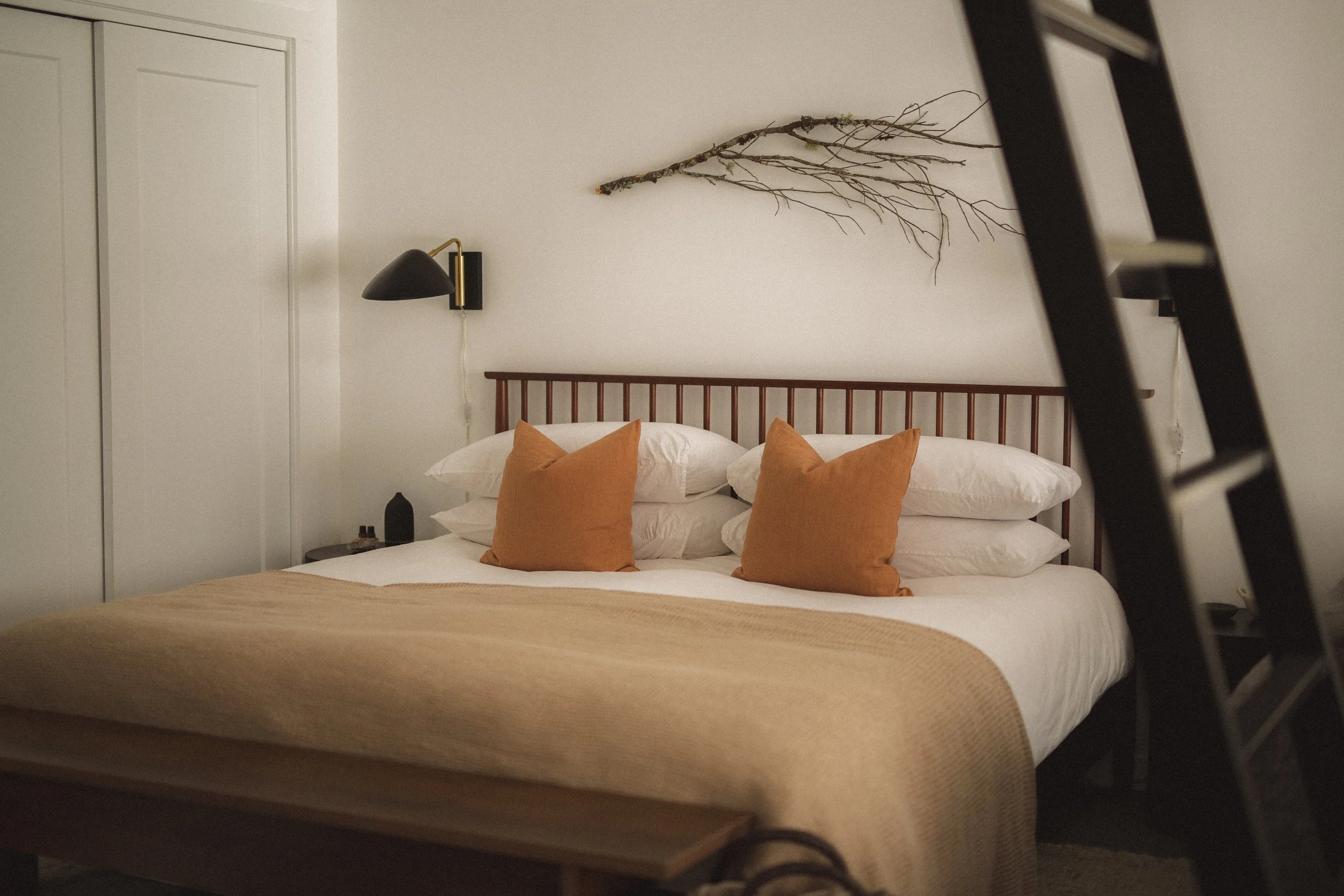 A neatly made bed with white sheets and beige blanket, four white pillows, and two rust-colored throw pillows in a bedroom with a black wall-mounted lamp, a twig decoration on the wall, a black ladder partially visible on the right side, and a white door on the left.