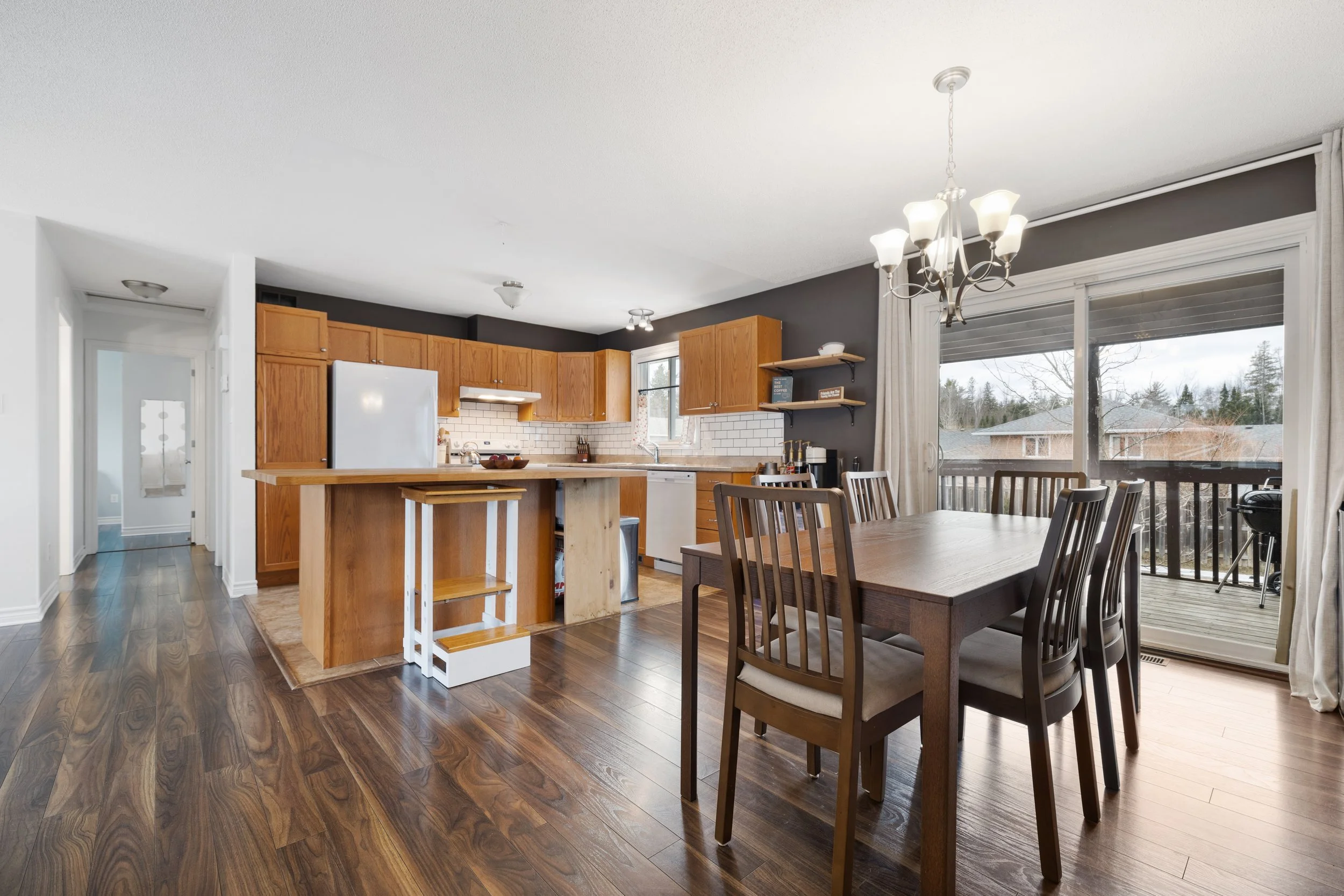 Open concept kitchen and dining area with wooden floors, a dining table with six chairs, and a sliding glass door leading to a balcony with a barbecue grill. Kitchen has wooden cabinets, white appliances, and a small island.