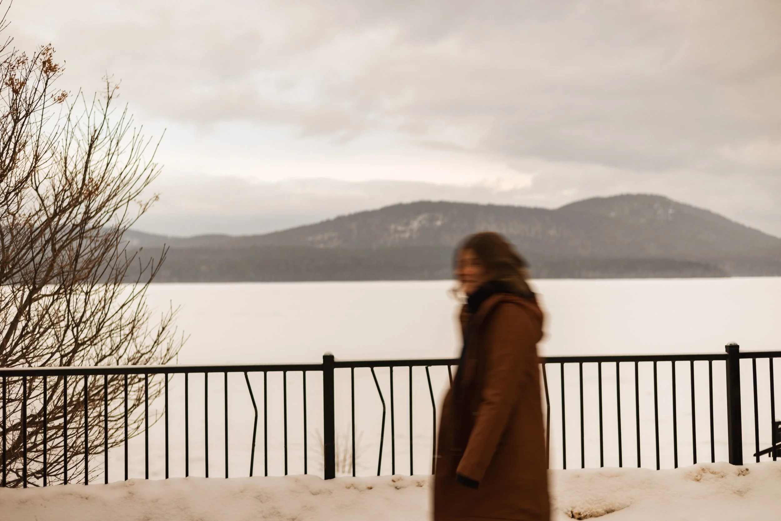 A person in a brown coat walking along a snow-covered path next to a black metal fence, with a lake and distant mountains under cloudy skies in the background.