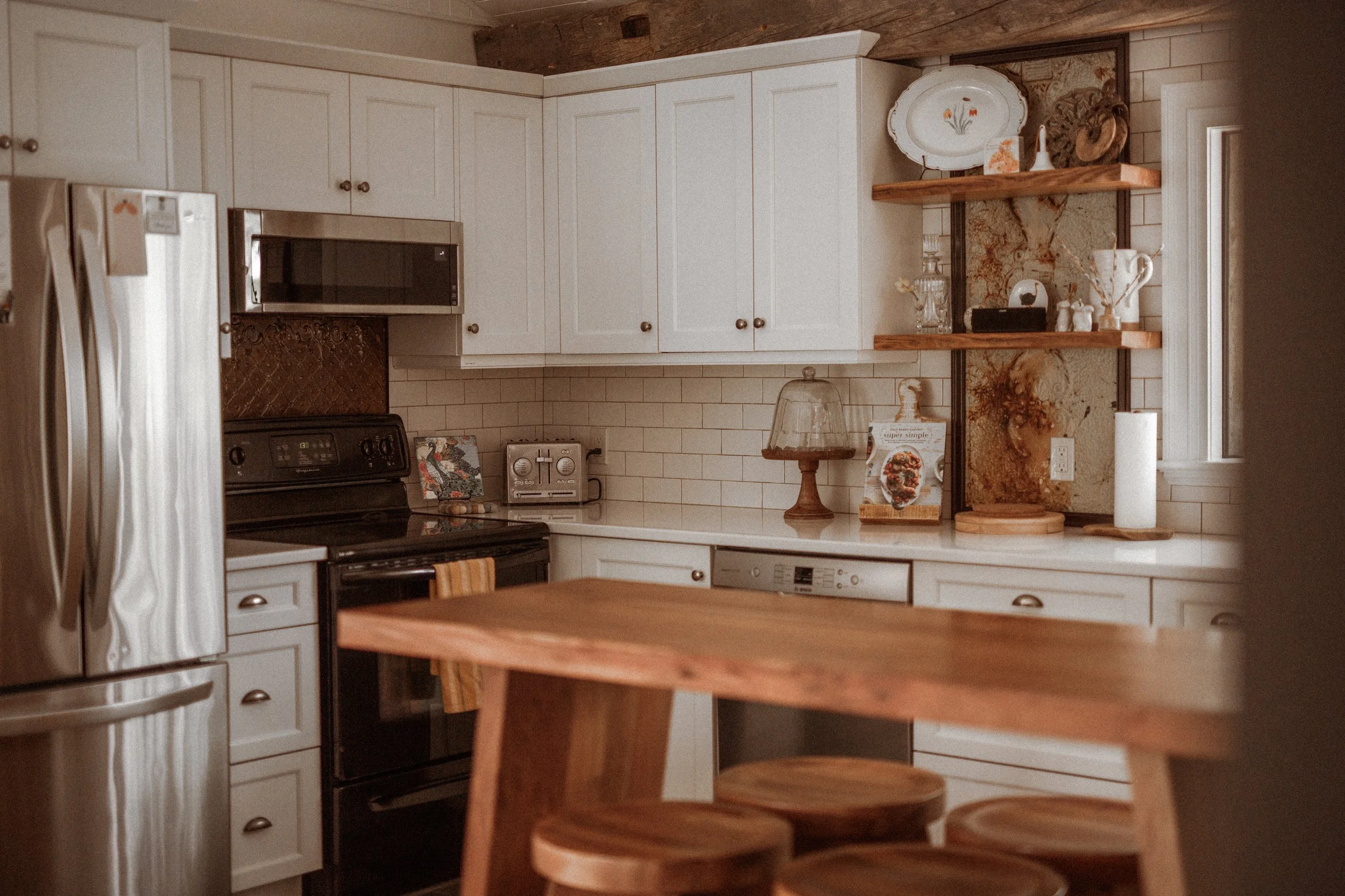 A cozy kitchen with white cabinets, a black stove, stainless steel refrigerator, and open wooden shelves displaying decorative dishes and items. A wooden table with matching stools is in the foreground, and a window lets in natural light.