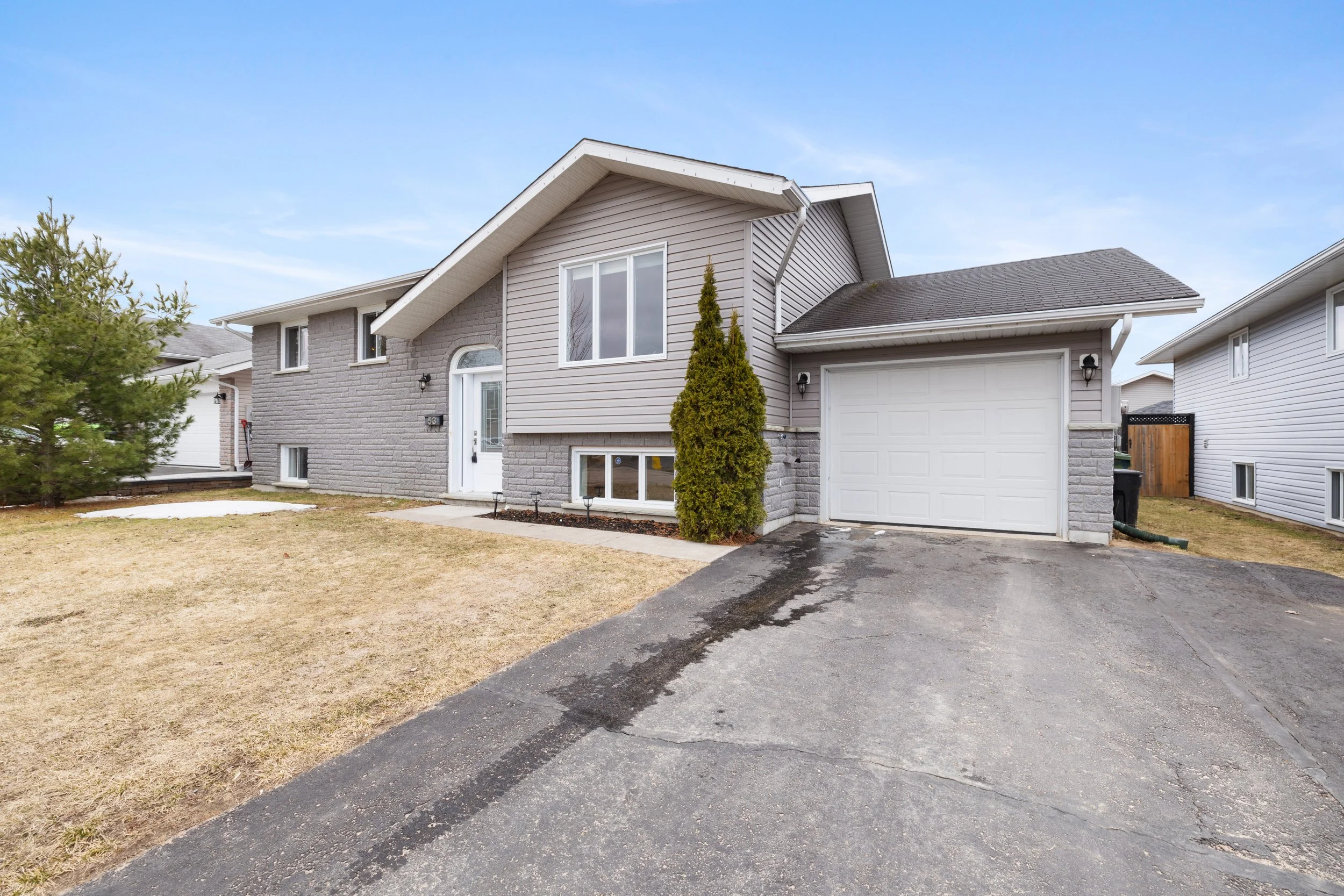 Front view of a two-story house with gray brick and vinyl siding, a white garage door, a small driveway, and a lawn with some patches of snow and bushes.