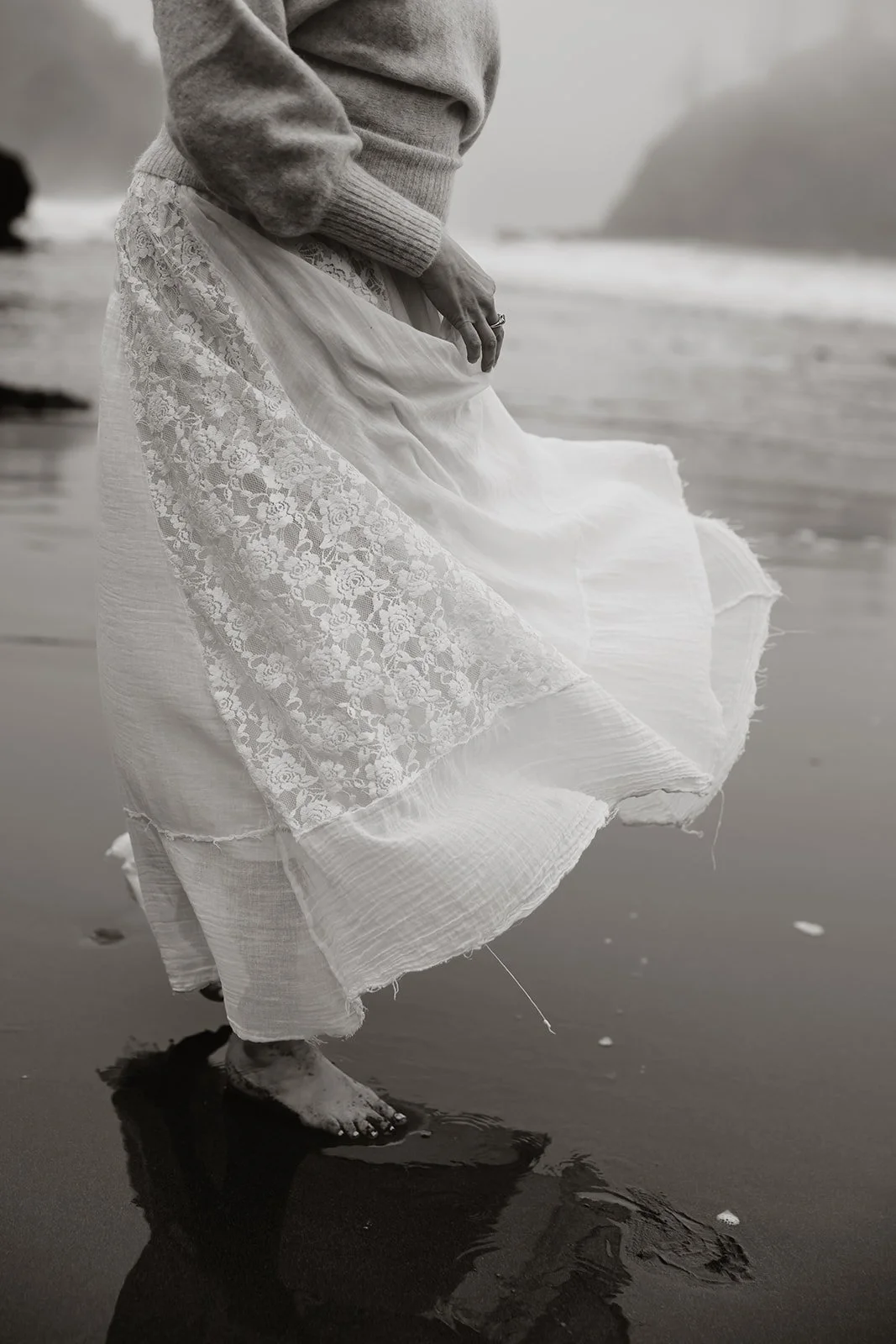 Person standing barefoot on wet sand, wearing a white lace skirt and gray sweater, near the shoreline with ocean and hills in the background.