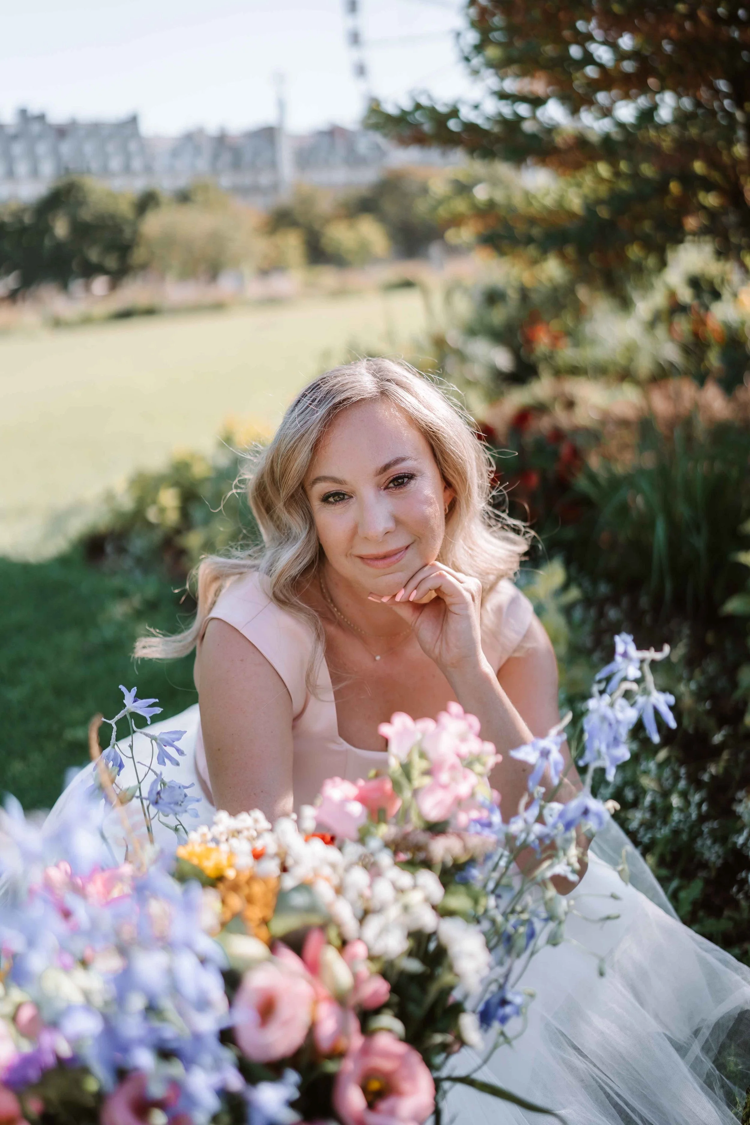 A woman with blonde hair in a light-colored dress sitting among colorful flowers in a park or garden during daytime.