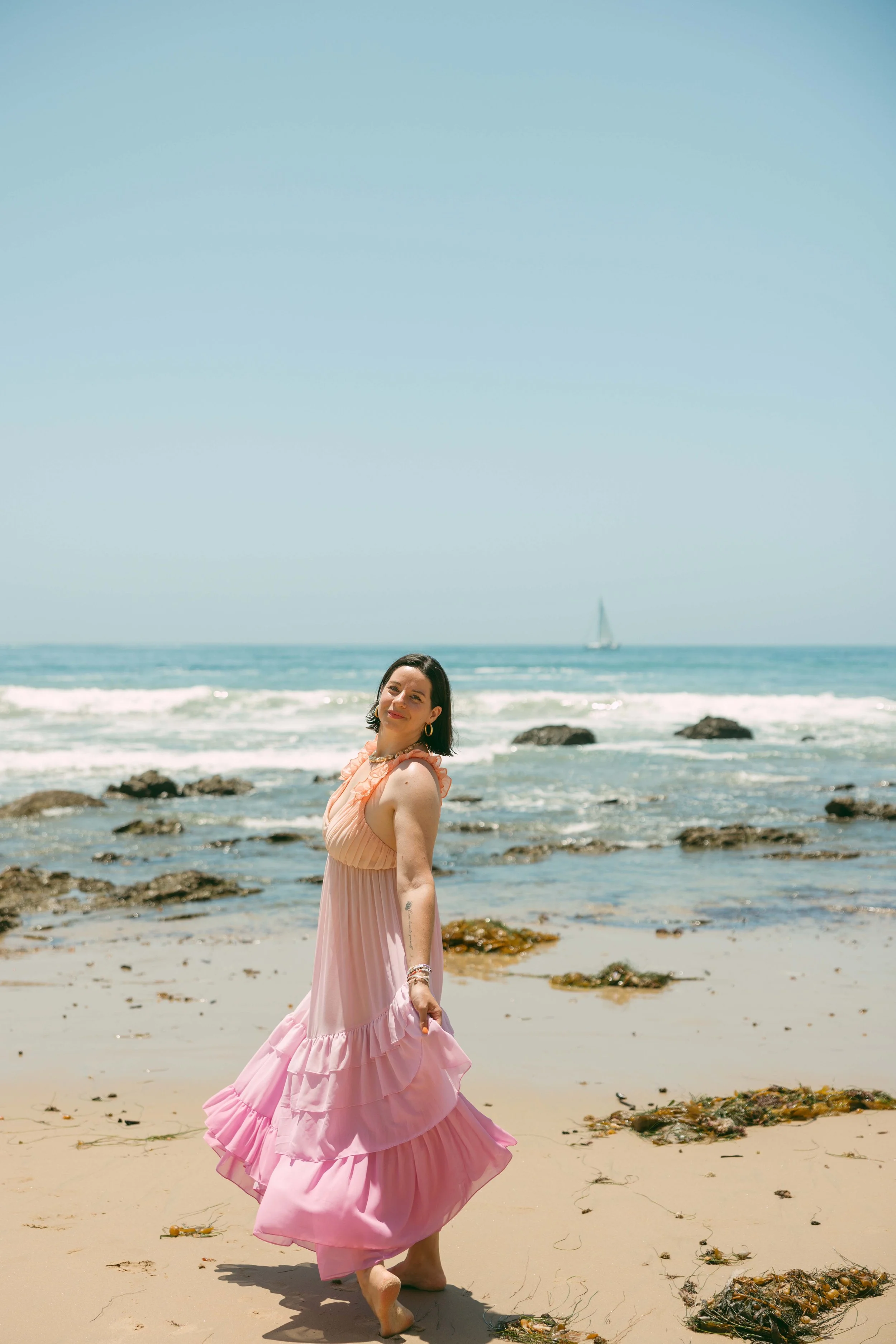 A woman in a pink flowing dress standing barefoot on a sandy beach with rocks, ocean waves, and a sailboat in the background.
