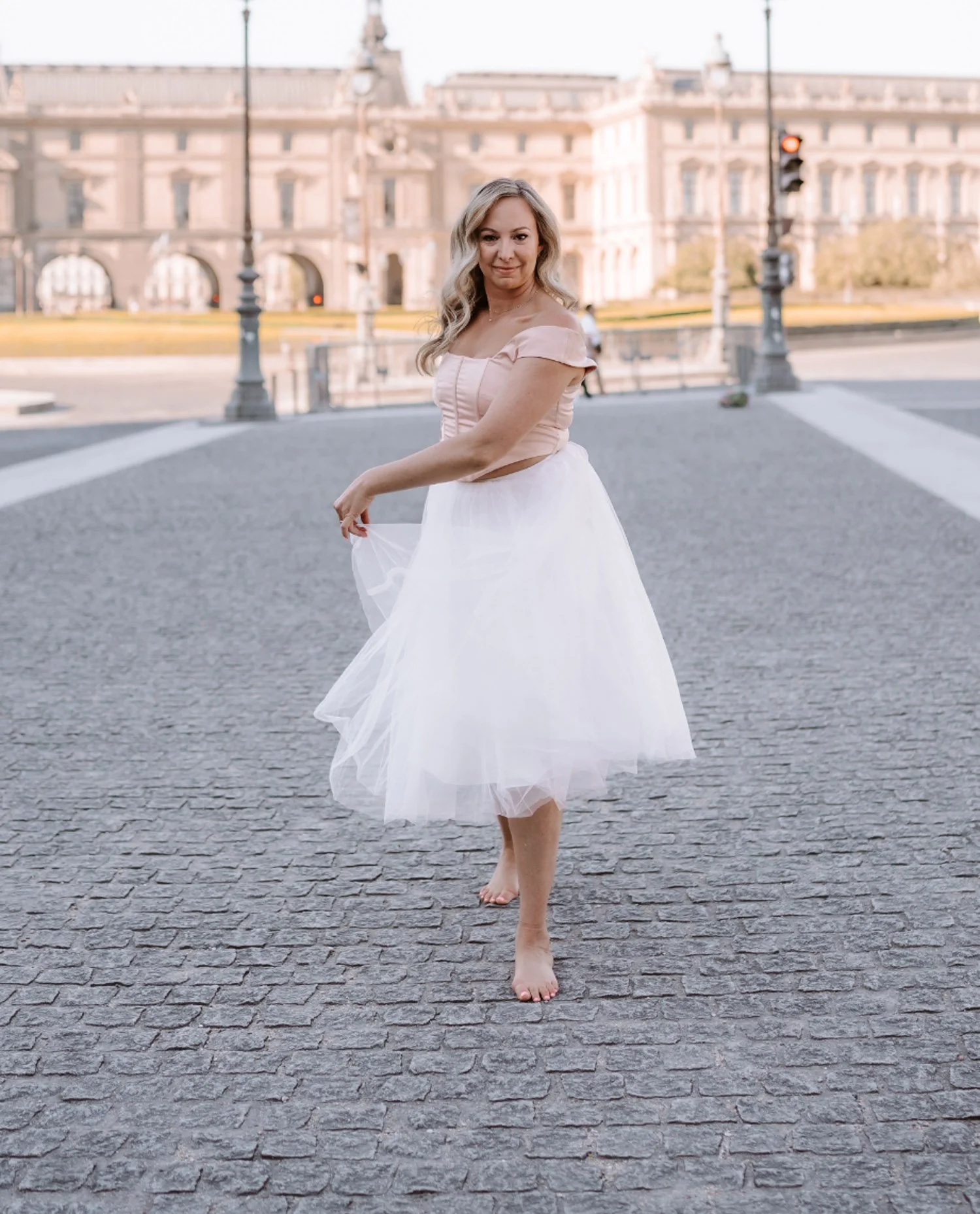 A woman standing barefoot on a cobblestone street, wearing a pink off-the-shoulder top and a white tulle skirt, with historic buildings in the background.