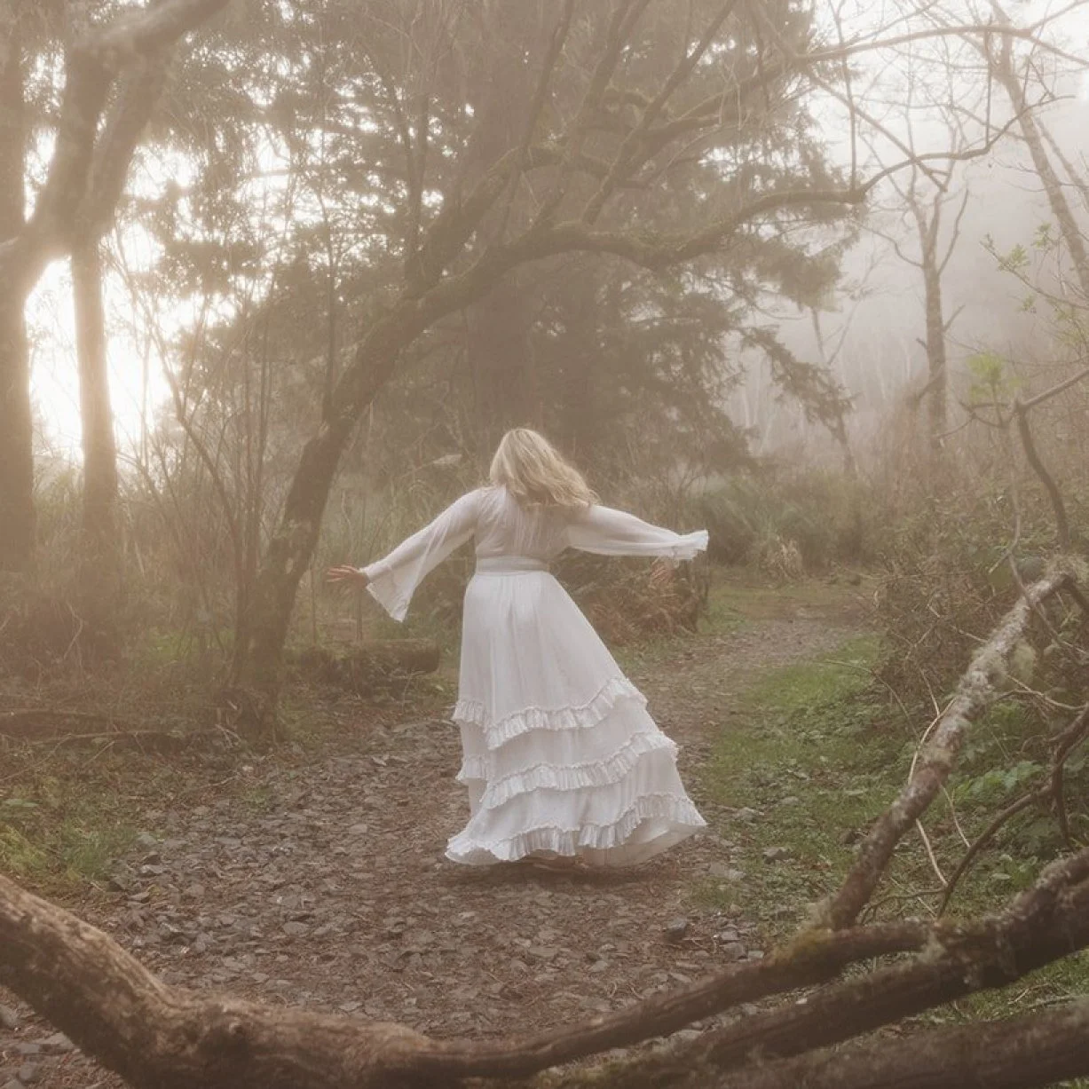A woman in a flowing white dress dancing on a wooded forest trail with misty, soft light filtering through the trees.