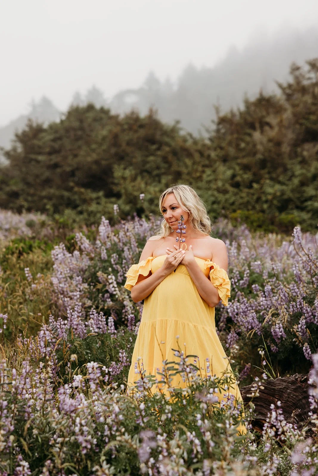 A woman in a yellow dress standing in a field of purple flowers, holding a flower close to her chest, with forested hills and fog in the background.