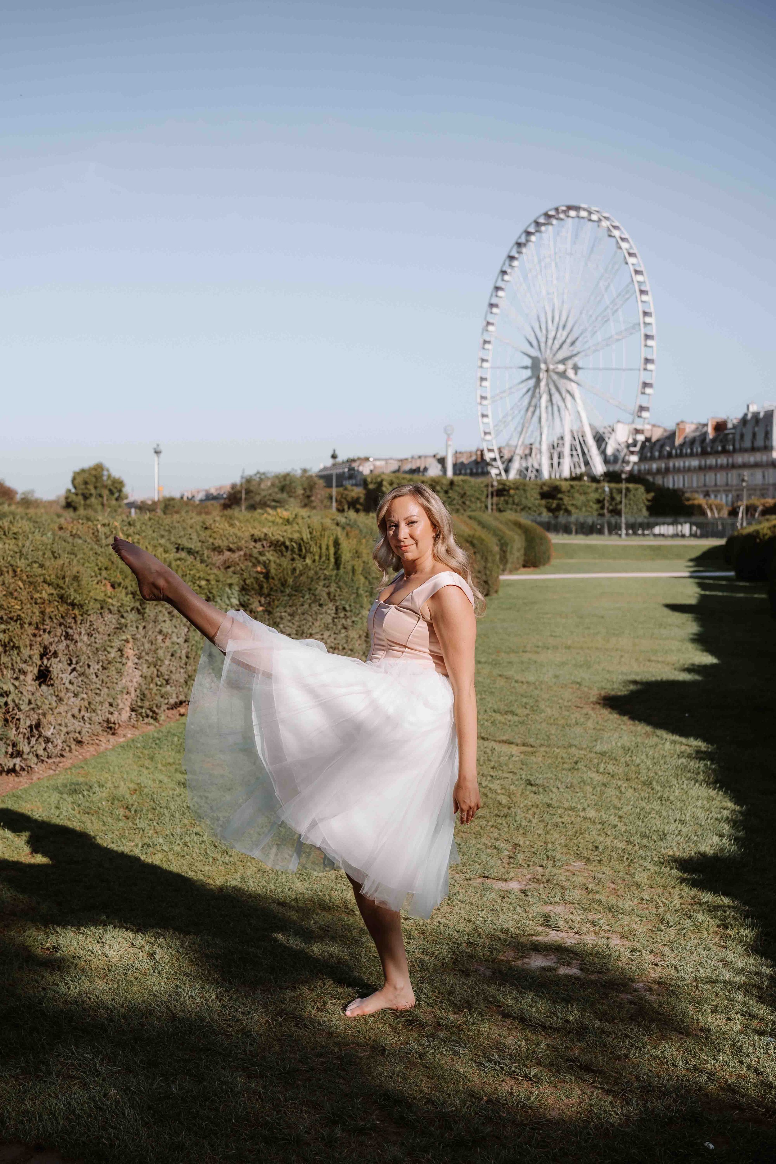 A woman in a light pink dress and tulle skirt performing a high kick in an outdoor park with a Ferris wheel in the background.