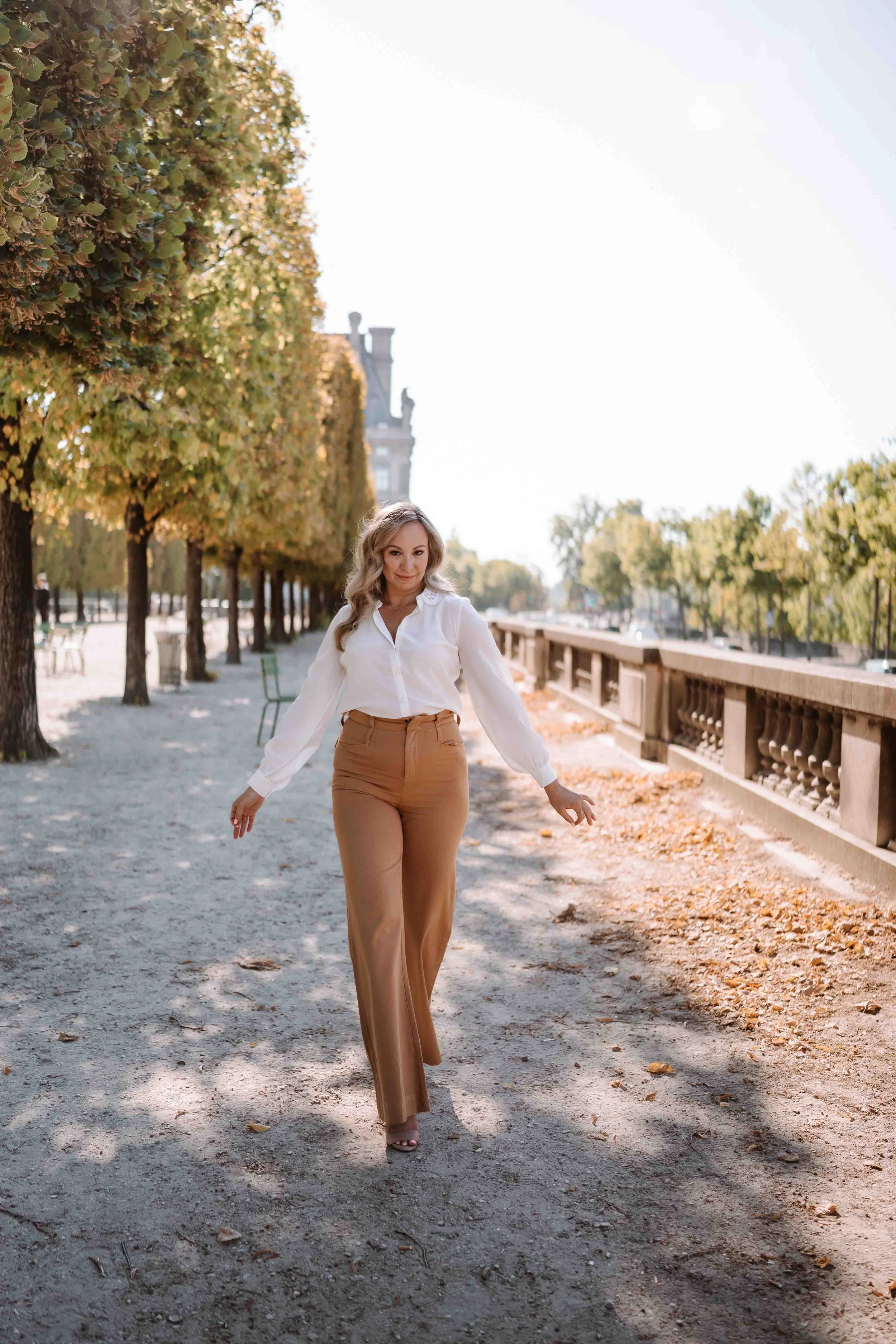 A woman walking on a dirt path lined with trees and a stone railing along a river, with a historic building in the background on a sunny day.