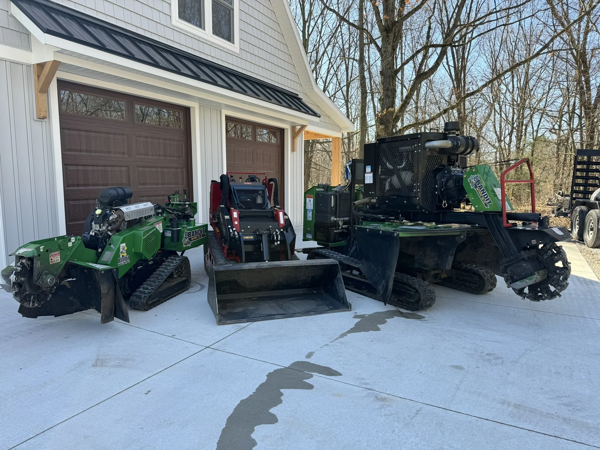 Three pieces of landscaping equipment and machinery parked on a concrete driveway in front of a house with two garage doors. The equipment includes a green tracked mower or trencher on the left, a small red mini skid-steer loader in the middle, and a larger green tracked mini excavator on the right.