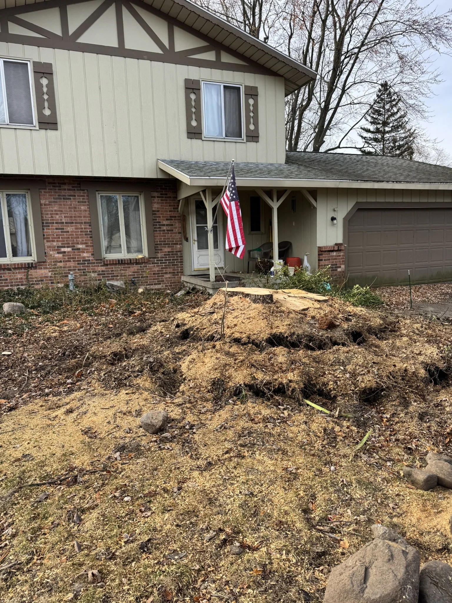 A front yard with a large tree stump and exposed roots, with a house in the background that has a beige and brick exterior, a small porch, and an American flag hanging from a pole.