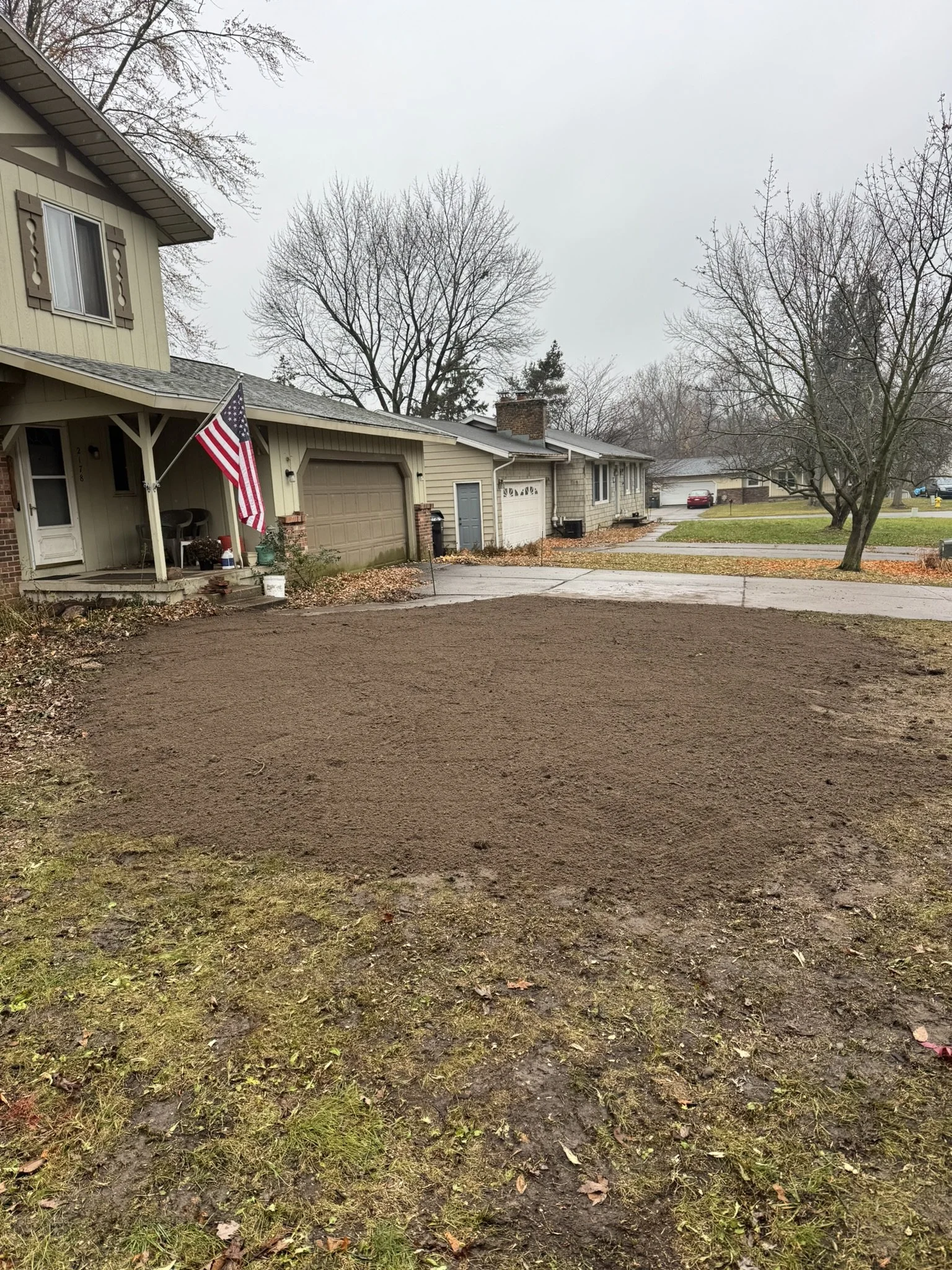 A suburban house with a front porch, American flag, and a newly leveled dirt yard in front. The house has a two-car garage and leafless trees are visible in the background on an overcast day.