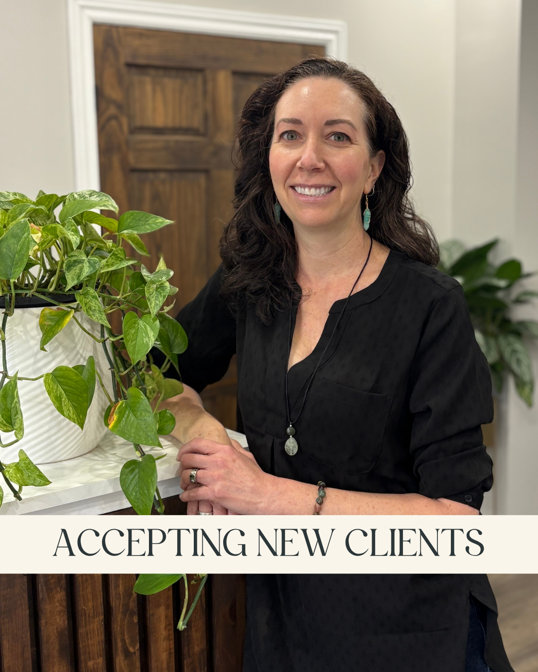 Deborah McEwan, MSW, RSW - A smiling woman with dark curly hair, wearing a black top and turquoise jewelry, and leaning on a wooden counter with a green leafy plant nearby and a caption that reads "Accepting New Clients."