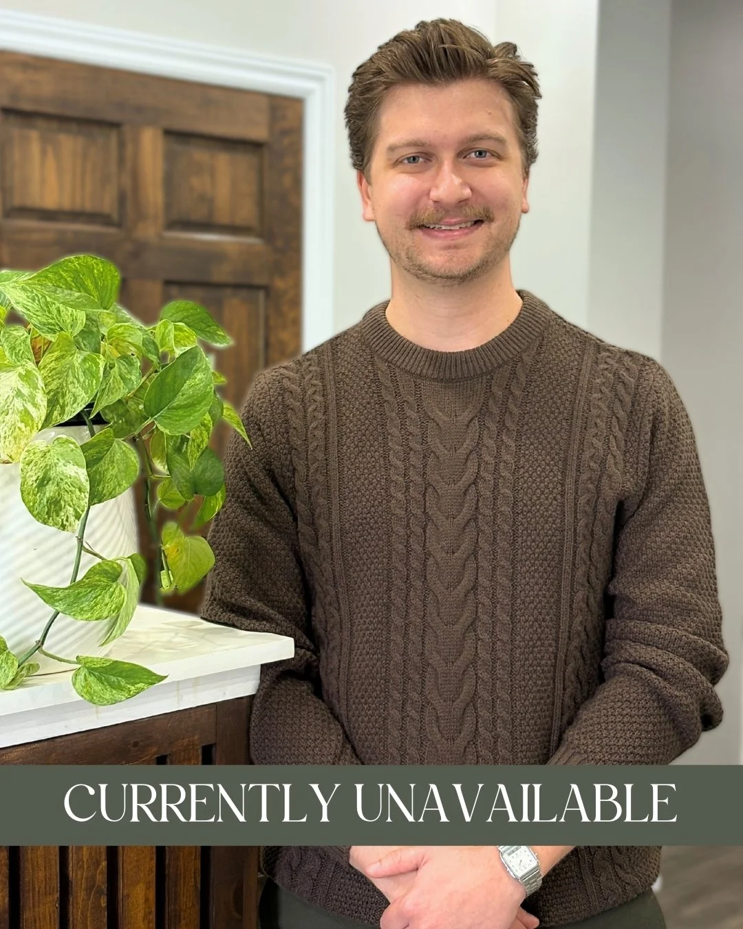 A man wearing a brown cable-knit sweater standing indoors next to a green leafy plant, with a wooden door in the background. The image has a "Currently Unavailable" overlay at the bottom.