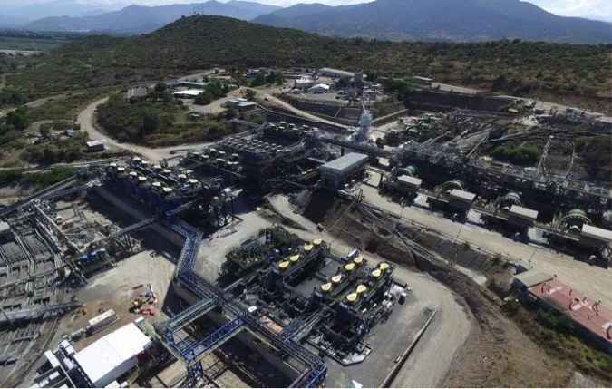 Aerial view of an industrial plant with machinery, pipelines, and equipment, surrounded by hilly terrain and mountains in the distance.