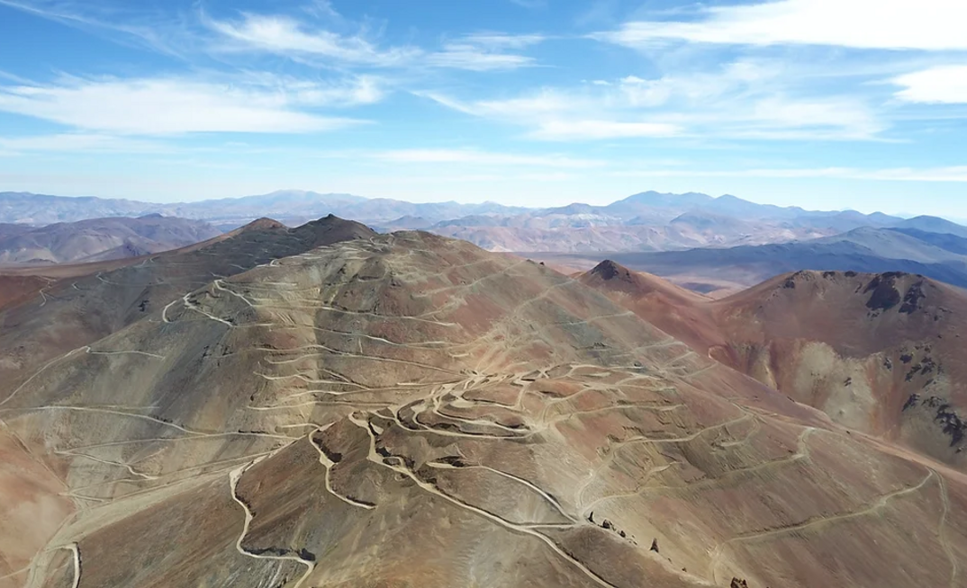 Aerial view of rugged mountains with winding dirt roads across the terrain under a blue sky with wispy clouds.