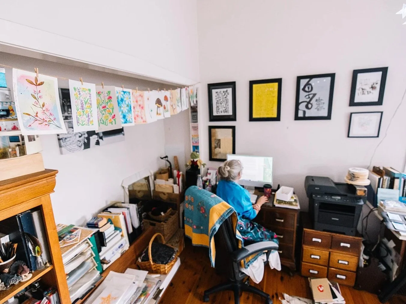 Liz working at her desk in a home office with colorful artwork hanging on a string above and framed pictures on the wall. There are books, papers, and office supplies around her.