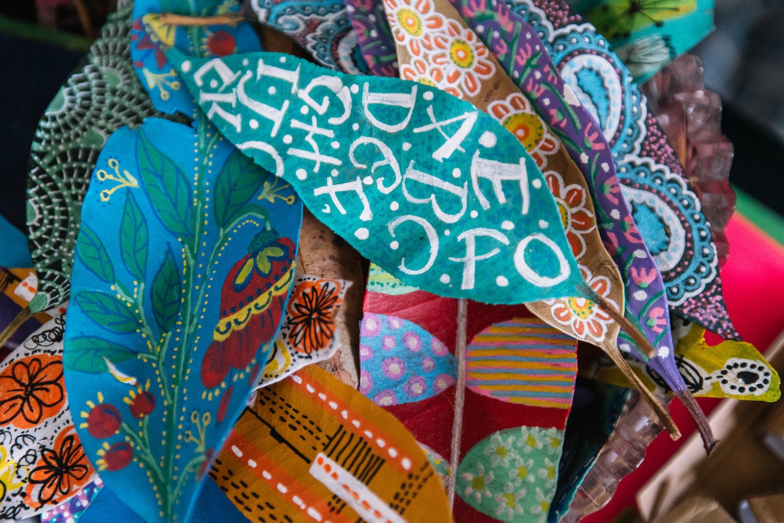 Colorful, hand-painted fabric leaves with various designs and patterns, including floral motifs, dots, and text, arranged in a bunch.