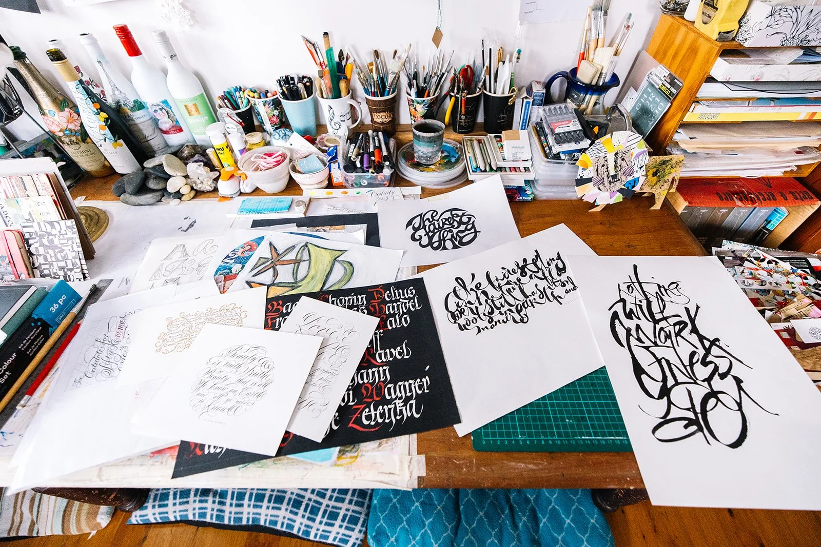 A cluttered art and calligraphy workspace with various papers, brushes, pens, and decorative bottles on a wooden table.