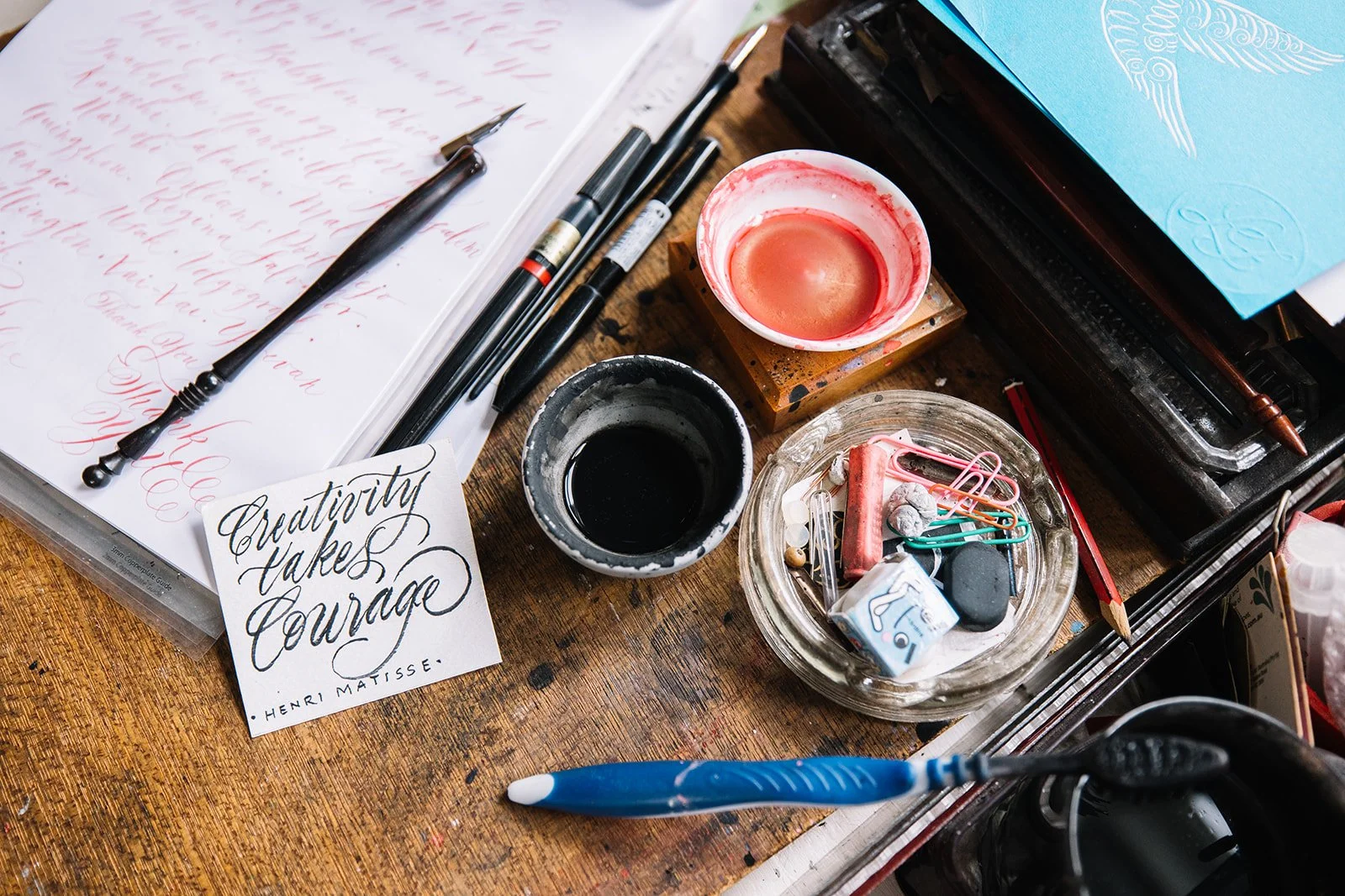 A cluttered desk with calligraphy supplies, ink jars, pens, paper, paper clips, and a card with a quote by Henri Matisse reading "Creativity Takes Courage."