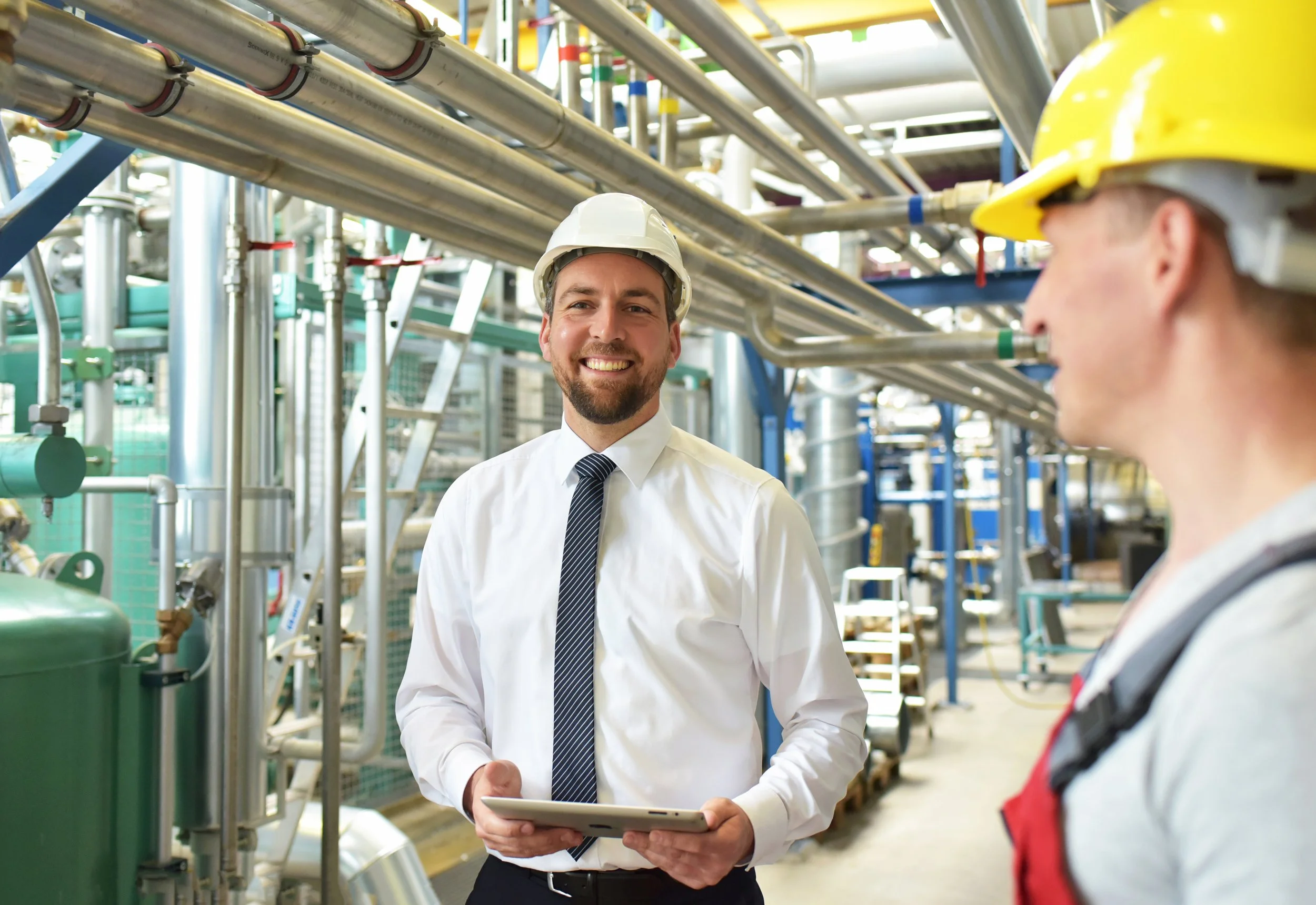 Two men in safety helmets, one in business attire and the other in work clothes, conversing in an industrial setting with pipes and machinery.