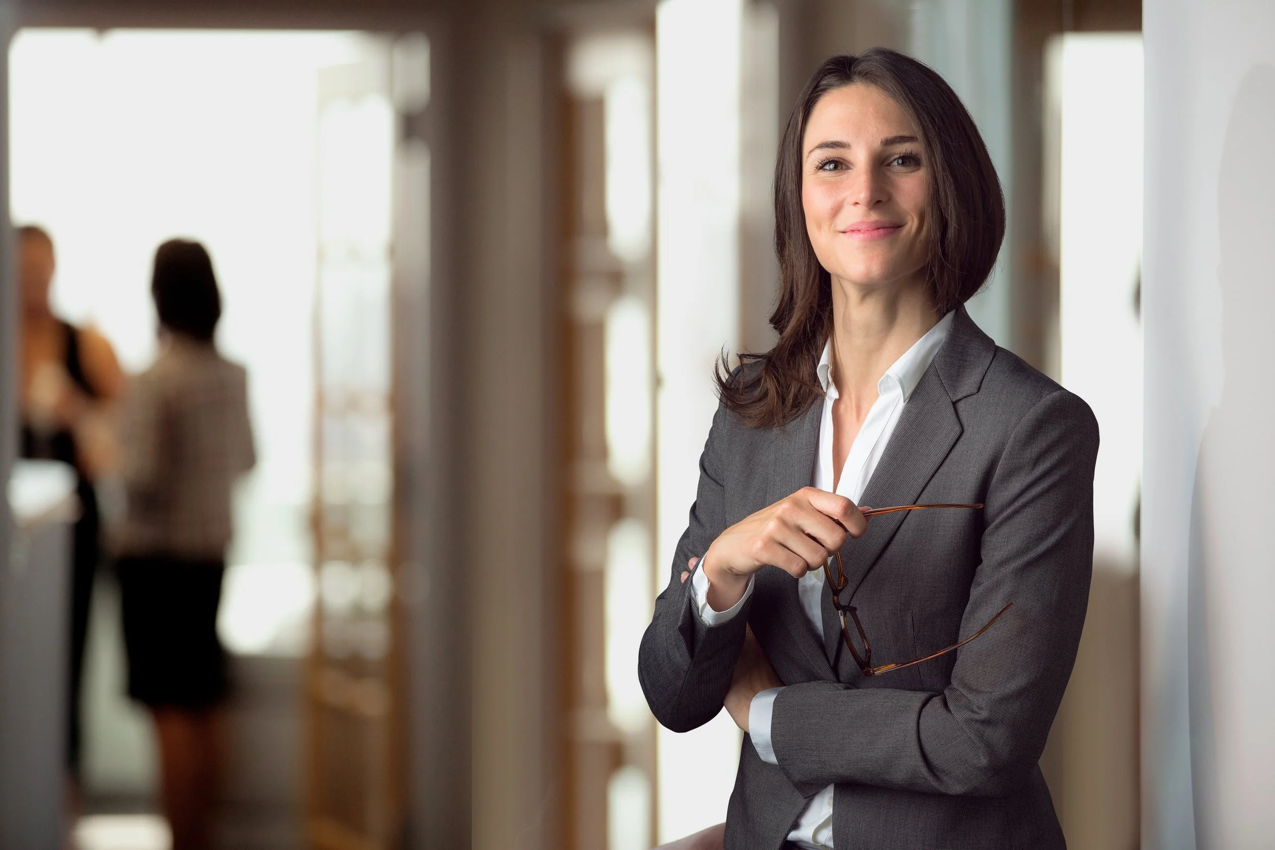 A professional woman in a gray suit holding glasses and smiling in an office corridor.