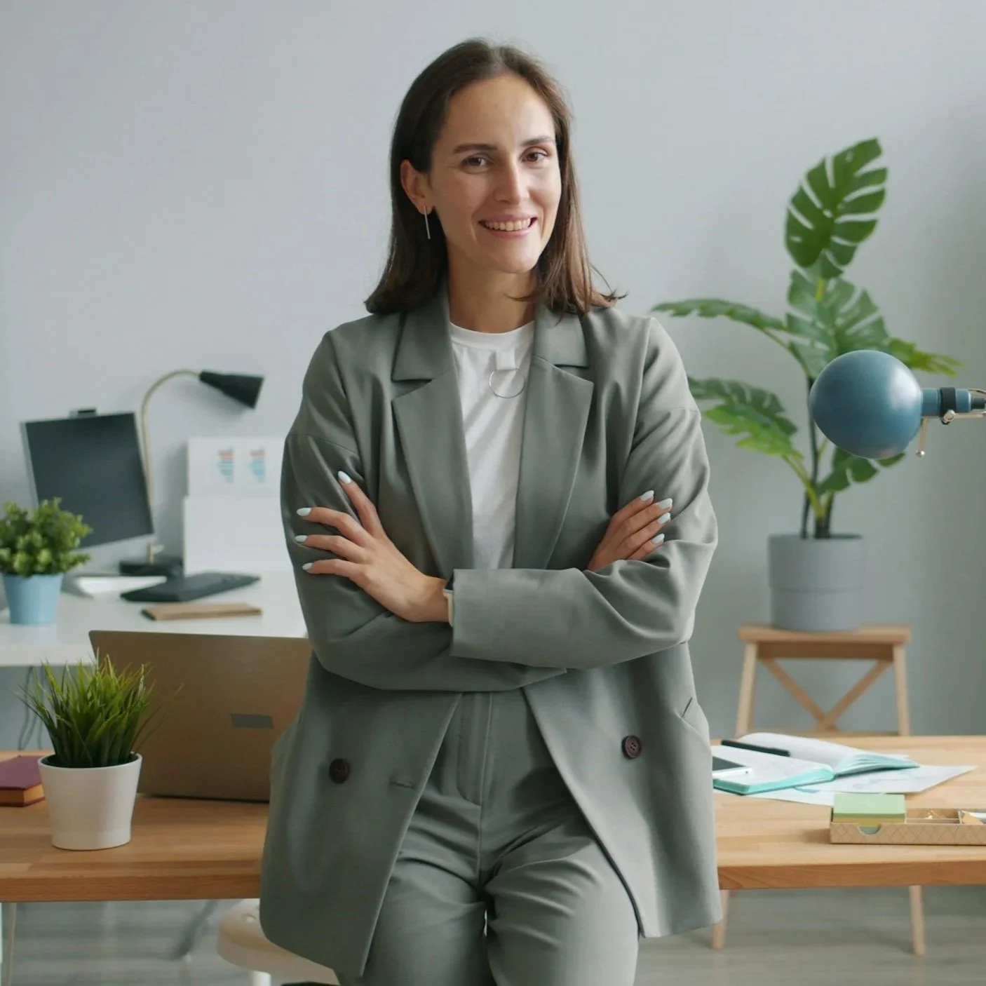 A woman with brown hair, wearing a gray blazer and pants, standing in an office with arms crossed, smiling at the camera.
