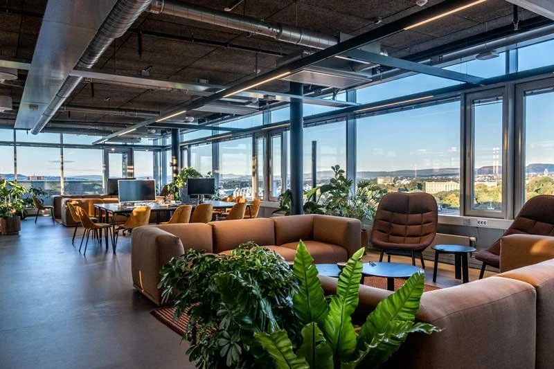 Innendørs kontorlokale i øverste etasje på Rebel med store vinduer og byutsikt, komfortable stoler, sofaer og planter.