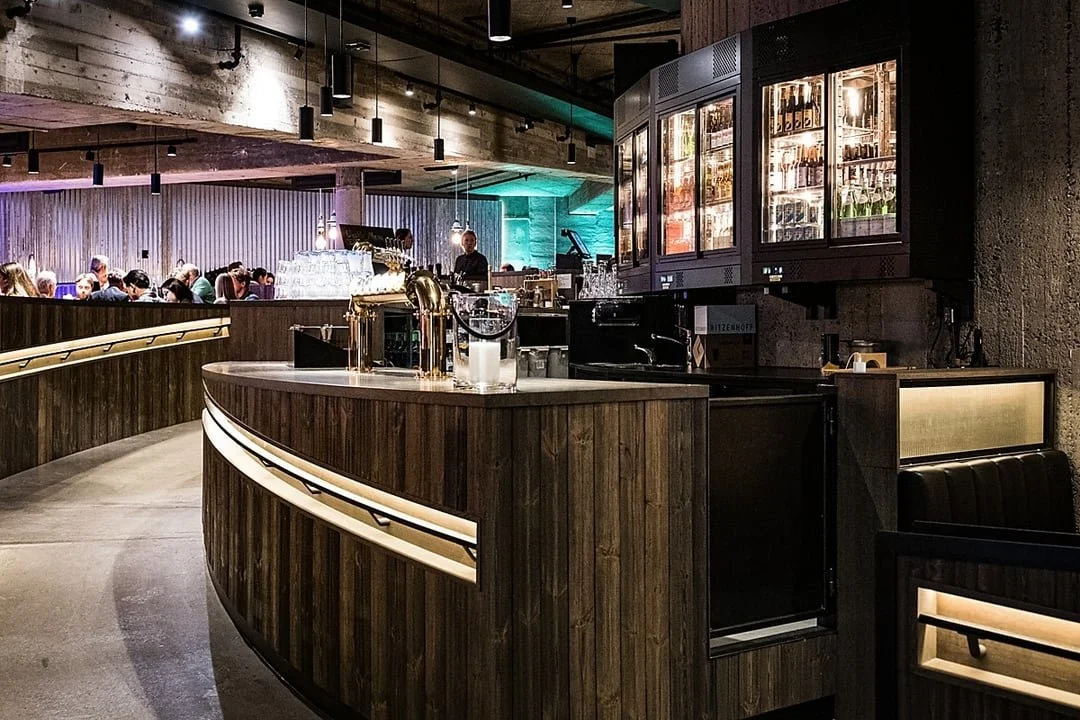 Interiør av en bar i en restaurant, med en buet tre- eller lamineringsbar, glassskap med drikkevarer, og mange mennesker som sitter i bakgrunnen.