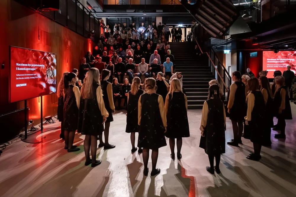 A choir performing in a modern auditorium with an audience seated on stairs. The event has red lighting and a screen displaying text in a foreign language.