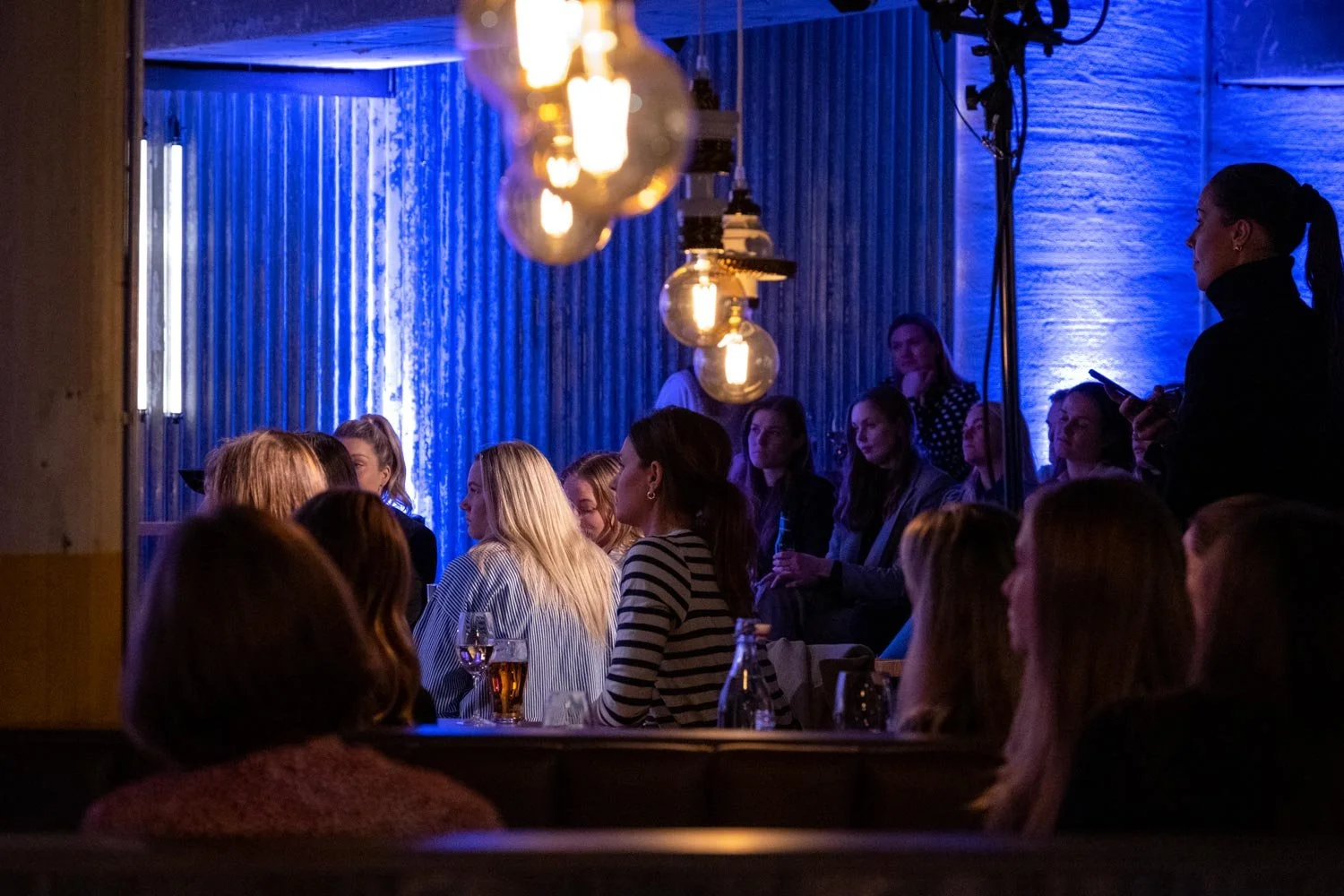 En gruppe mennesker sitter i et mørkt rom med blå belysning, og noen kvinner er i midten, mens en kvinne på høyre side står og bruker telefonen sin.