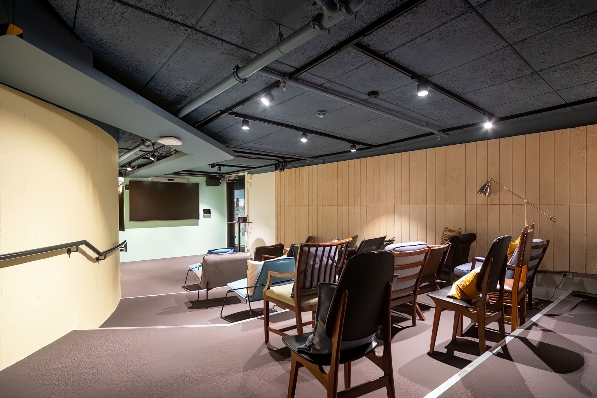 Modern conference room with wooden chairs and plush seating, large monitor, and adjustable lighting in an industrial-style space.