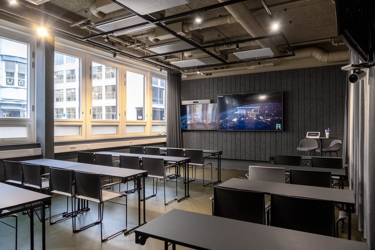 Modern classroom with desks, chairs, large windows, and a digital screen on the wall.
