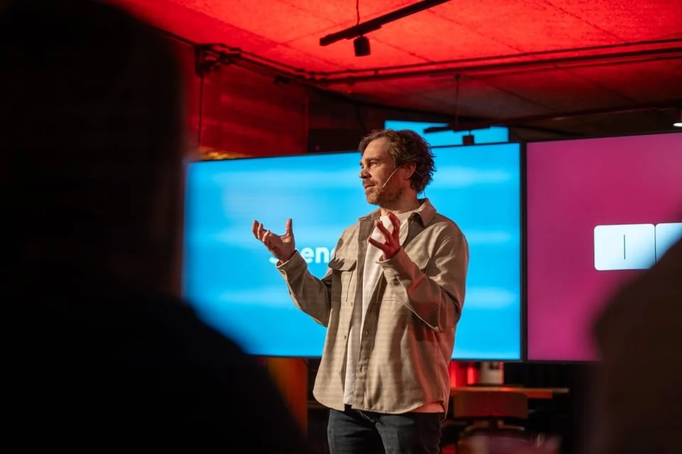 En mann som holder en tale eller presentasjon foran et publikum i et rom med store skjermer i bakgrunnen.