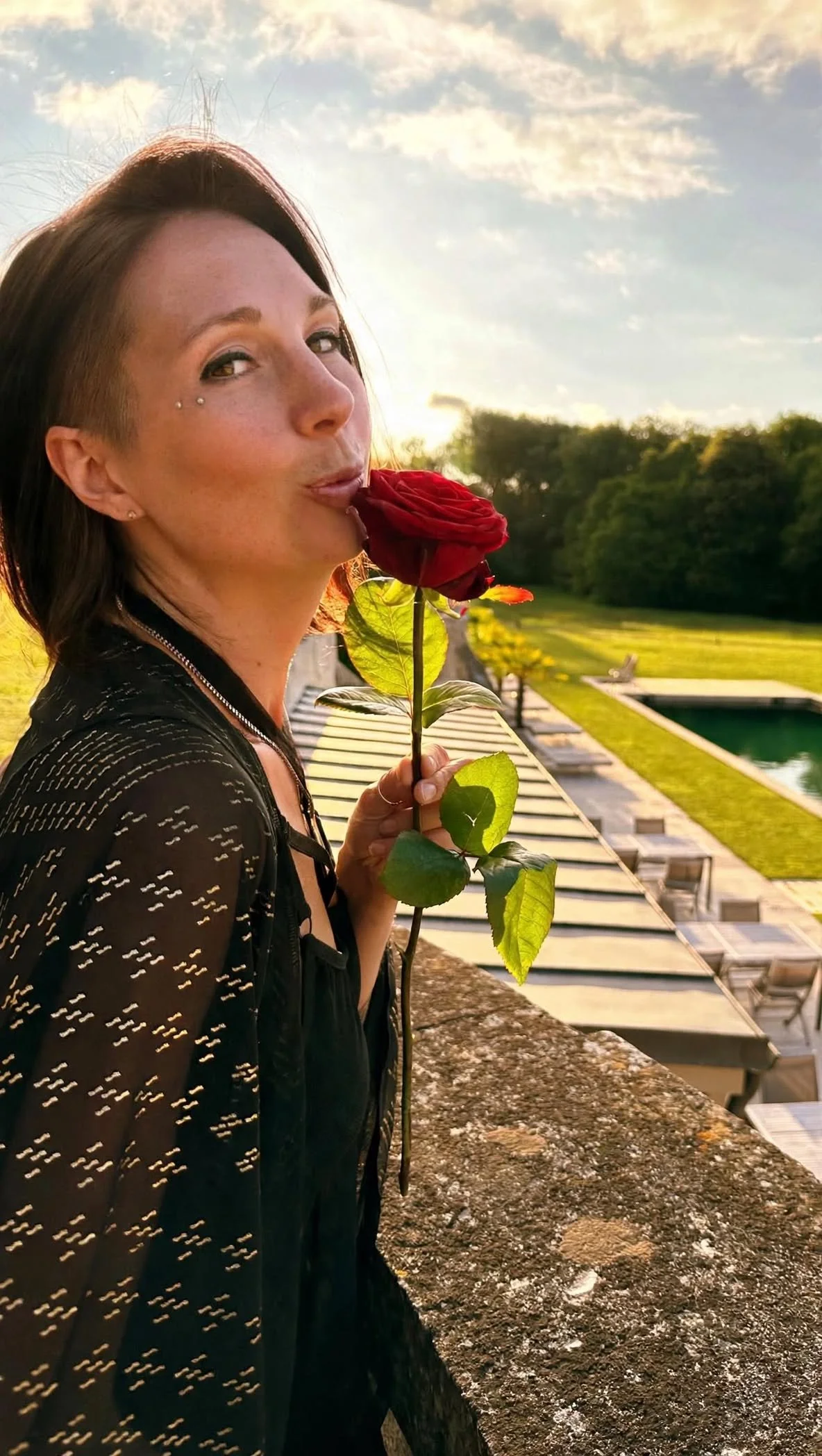 A woman with short brown hair and a diamond-studded eye piercing holds a red rose near her face. She is outdoors during sunset, with a green landscape, pool, and lounge chairs in the background.