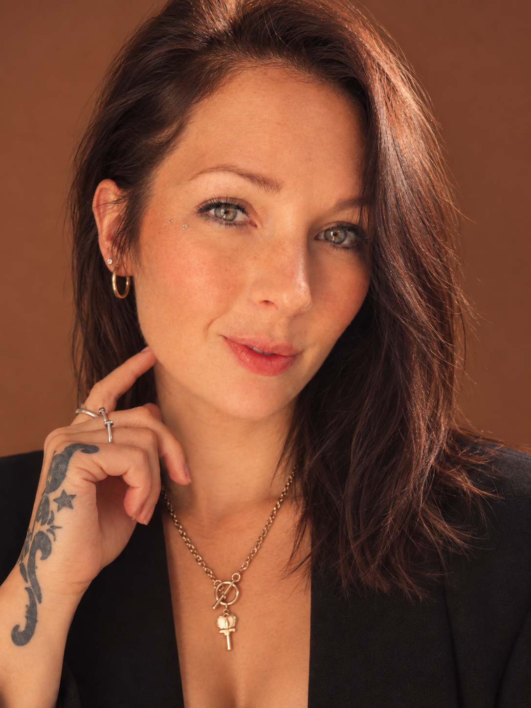 A woman with shoulder-length dark brown hair, light skin, and striking blue eyes, wearing gold hoop earrings, gold necklaces with a key and a round pendant, and showing a tattoo on her left hand and wrist. She is touching her cheek with her left hand and has a subtle smile.