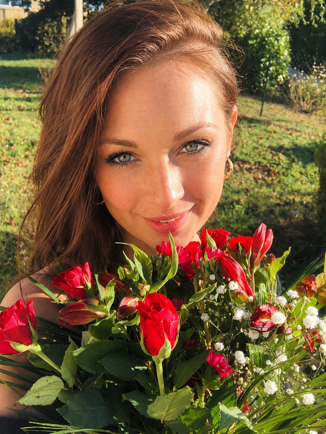 A young woman with reddish-brown hair and light-colored eyes holding a bouquet of red and white flowers outdoors on a sunny day.