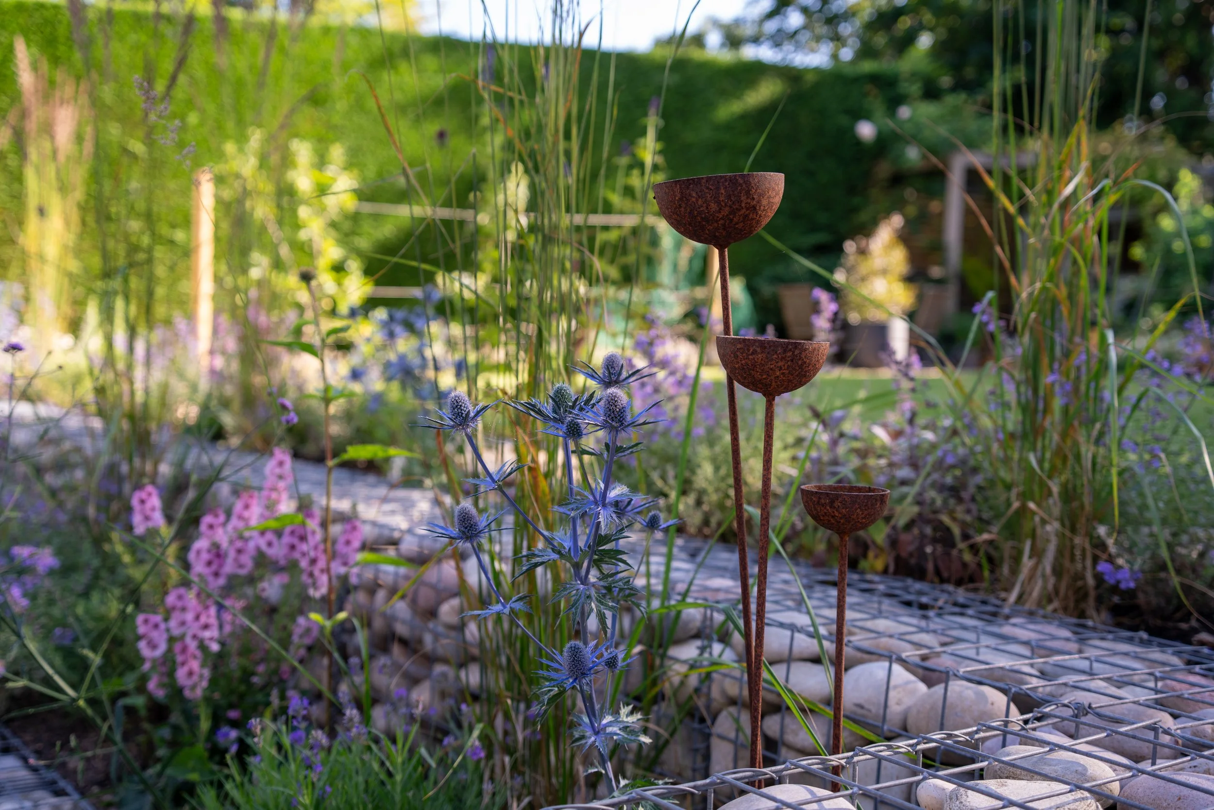 Rusty metal sculptures in a garden surrounded by purple and pink flowers, green foliage, and a pathway with rocks.