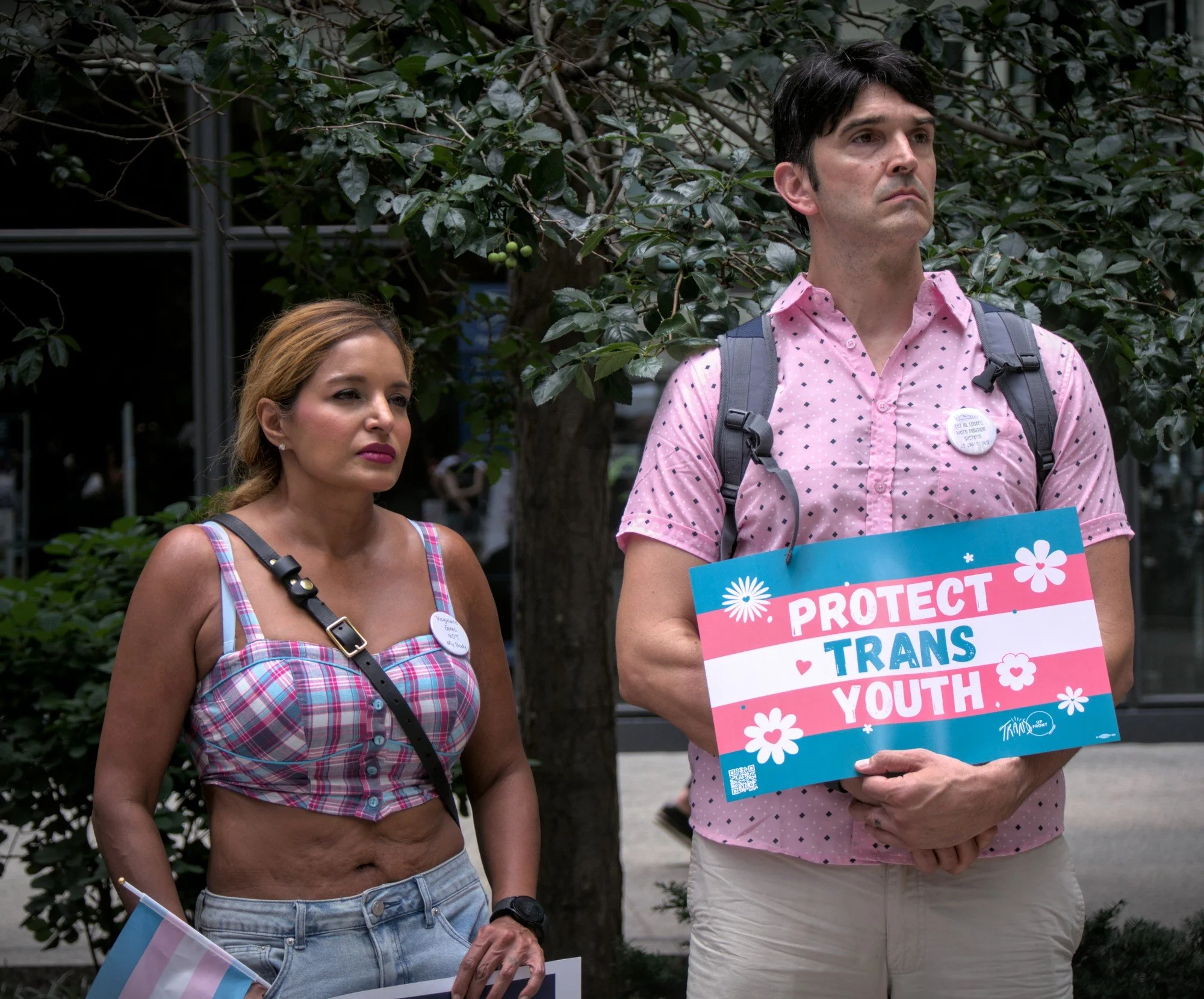  Two figures stand in front of a tree in the plaza. The one on the left is a shorter femme with dyed blonde hair, a trans pride colored plaid tank, and blue jean shorts. On the right, a taller and very dignified looking masc-appearing person wears a pink shirt with a button that reads 