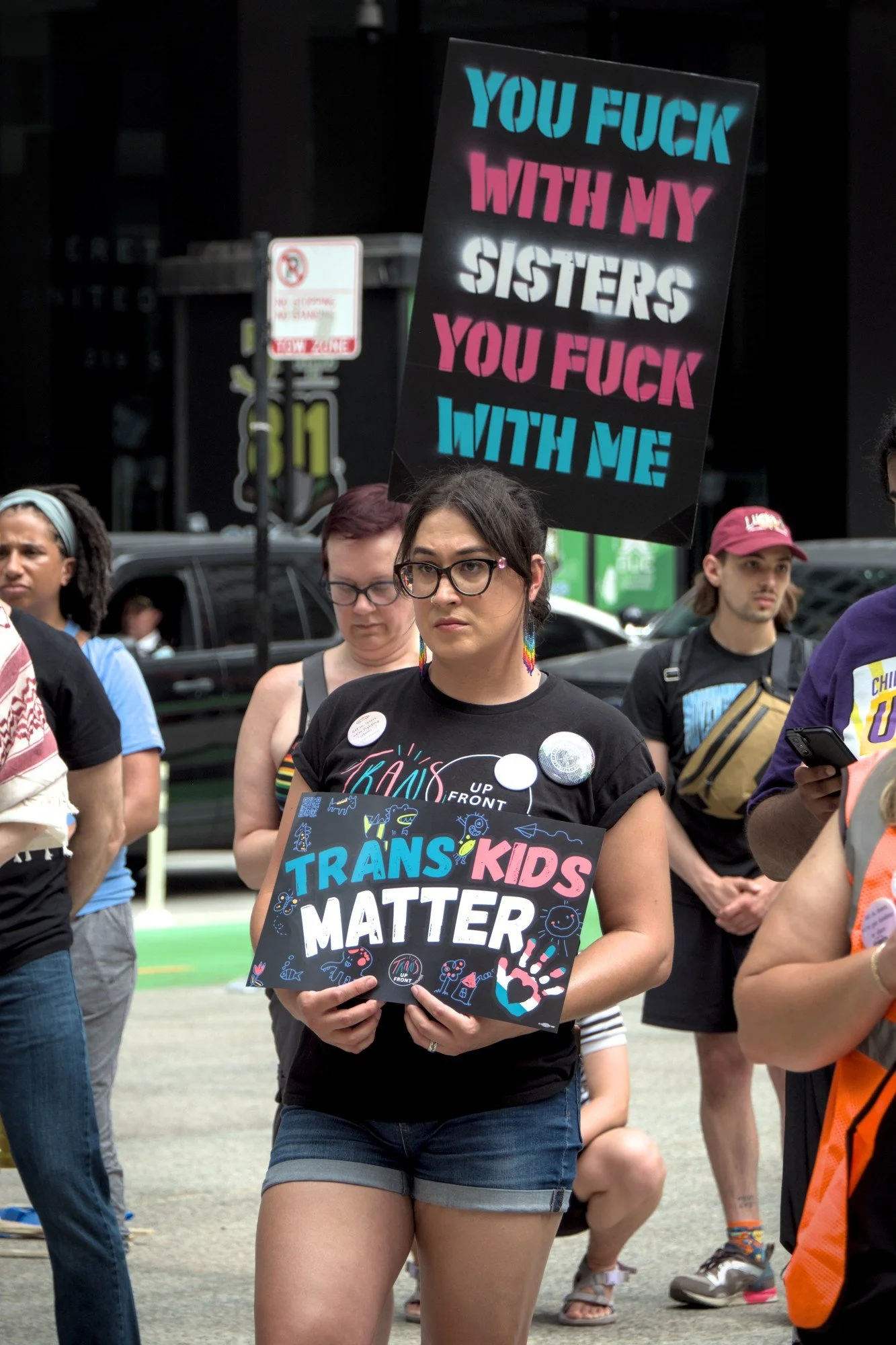   Dr. Corey Lascano, wearing jean shorts, a Trans Up Front shirt, and thick rimmed glasses stands with her shoulders back, holding a sign reading 