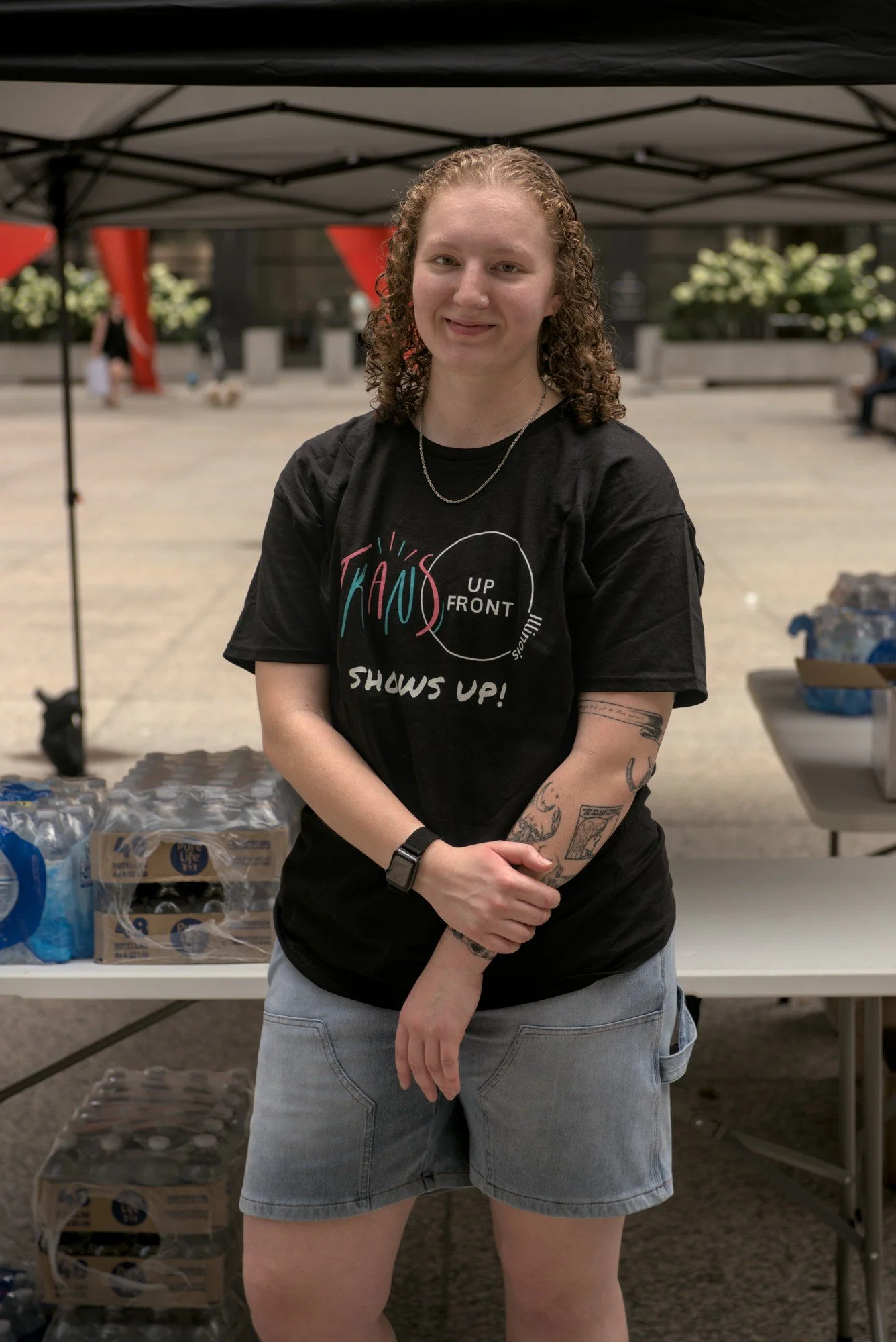   A portrait of Starr, a tattooed person with curly hair and a simple chain necklace wearing a shirt which reads TRANS UP FRONT SHOWS UP, standing in front of an all-set-up tent with cases of water on a folding table.  