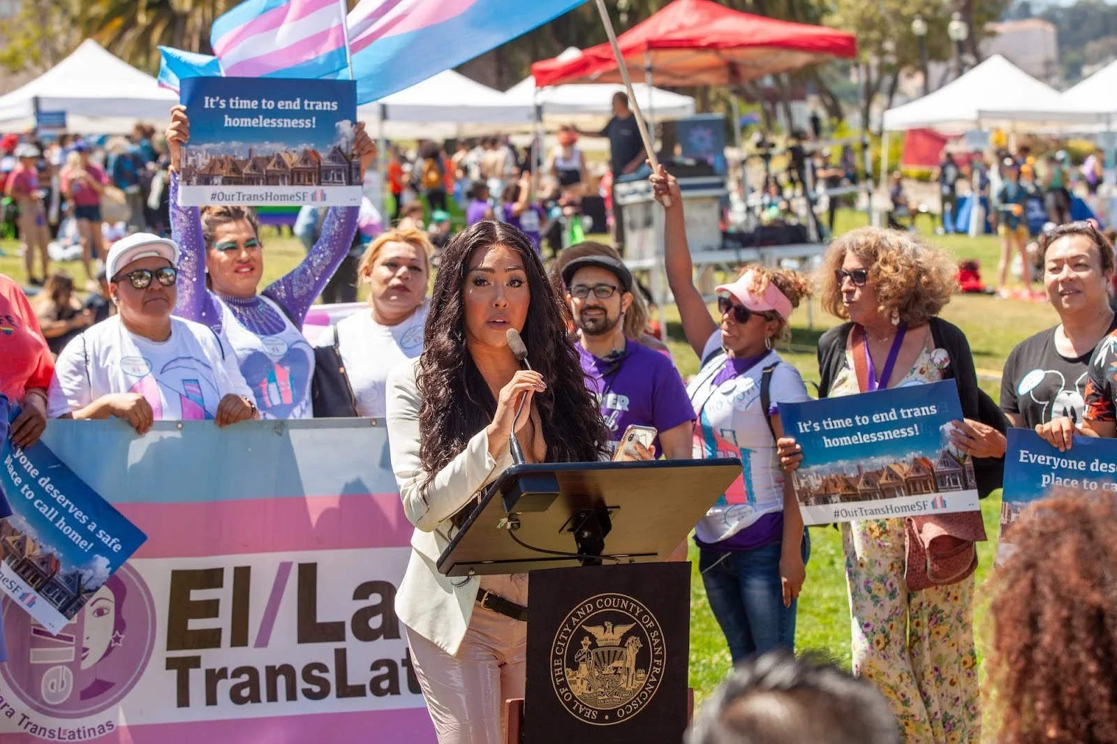    Melanie Ampon   , then member of the SF Human Rights Commission, speaks at a press conference at the    San Francisco Trans March   , June 28, 2019, surrounded by people holding signs reading “It’s time to end trans homelessness! #OurTransHomeSF” and a banner for    El/La Para TransaLatinas   .    Photo    licensed CC BY-SA 4.0.   