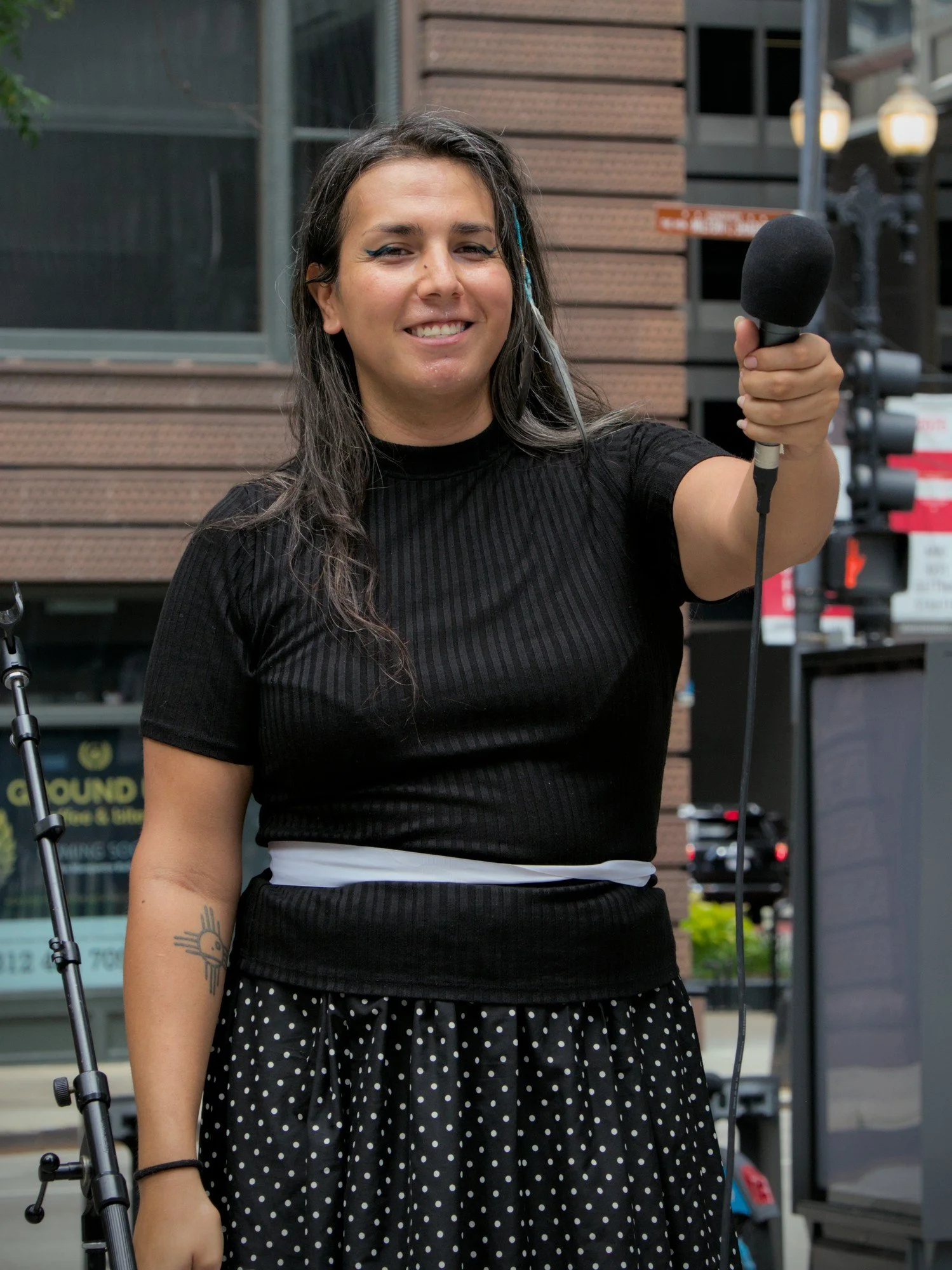   Another portrait of Albie Gutierrez, a brown-haired femme with a blue beaded streak tied into her hair wearing a skirt and black shirt, holding the microphone towards the crowd and smiling.  
