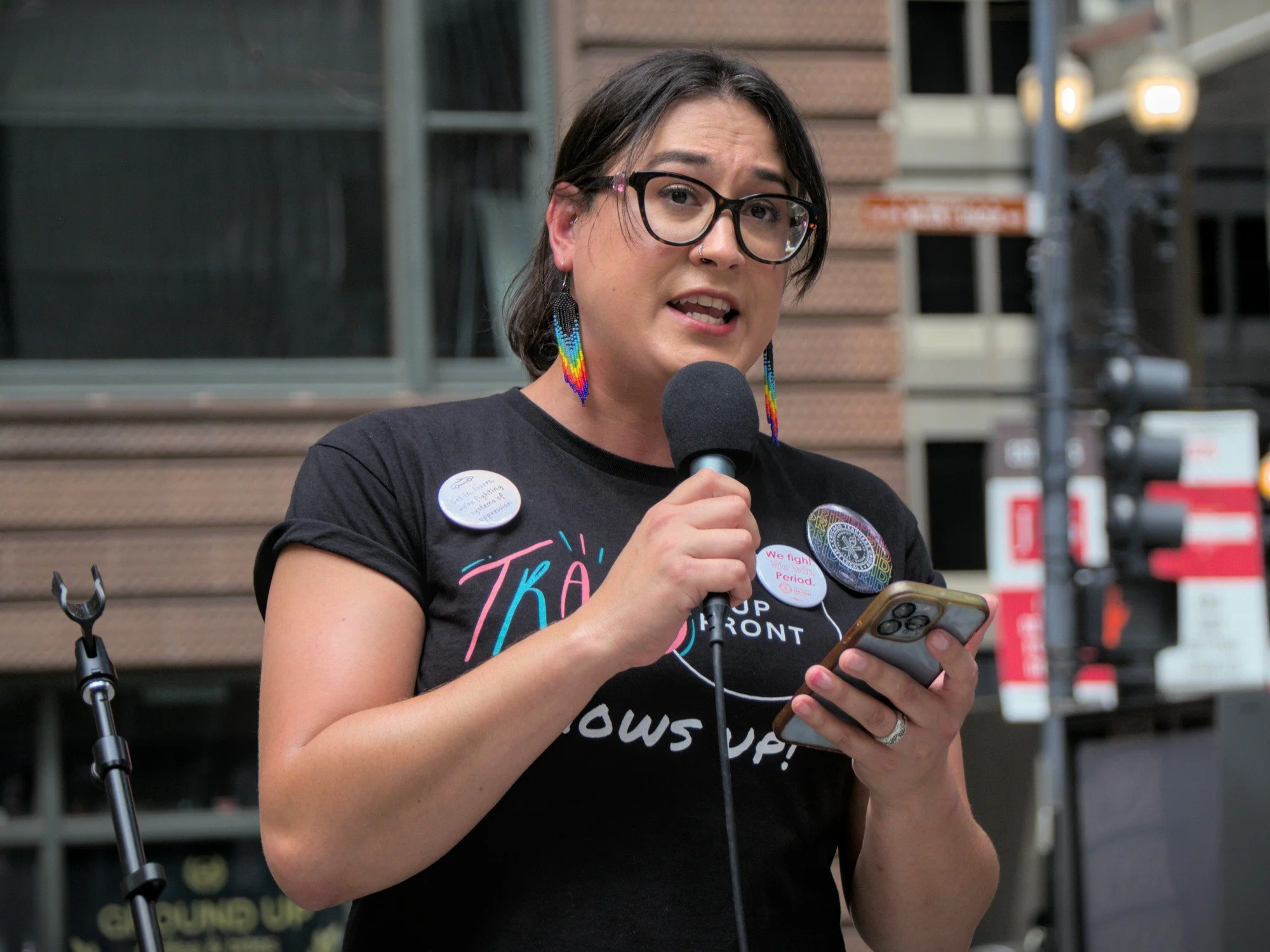  A portrait of Corey Lascano, a femme wearing glasses and beaded earrings with their hair tied back in a pony tail, reads from an Iphone and speaks to the crowd. 