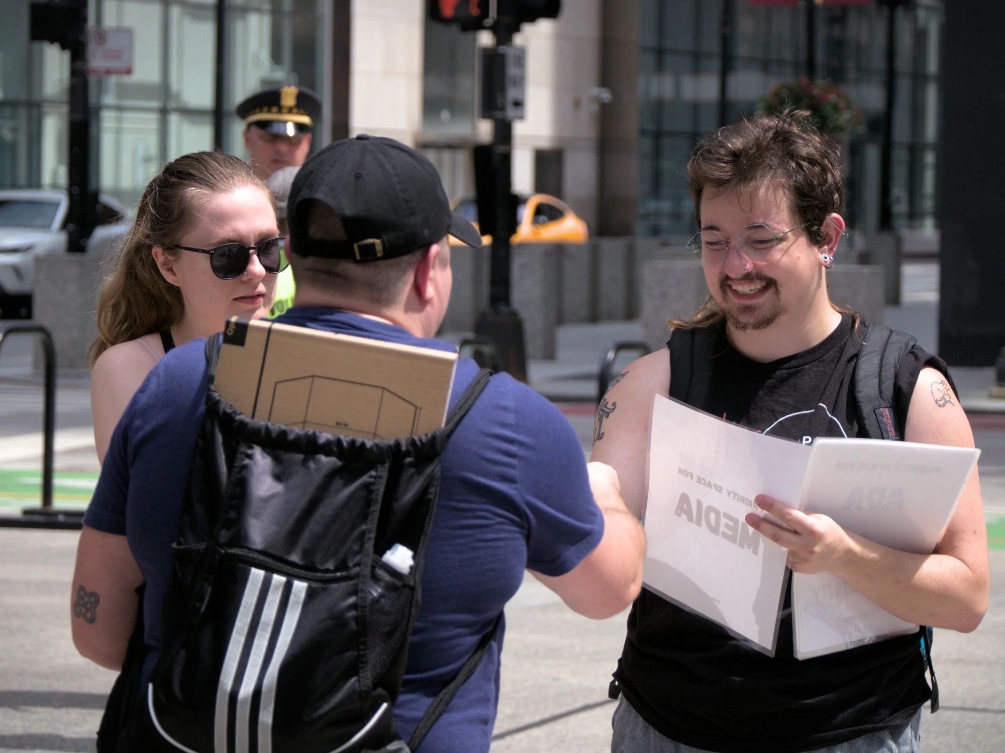   A smiling bearded masc wearing a TRANS UP FRONT tank hands out supplies and tape-marker signs to mark off sections of the plaza for media and ADA attendees.  