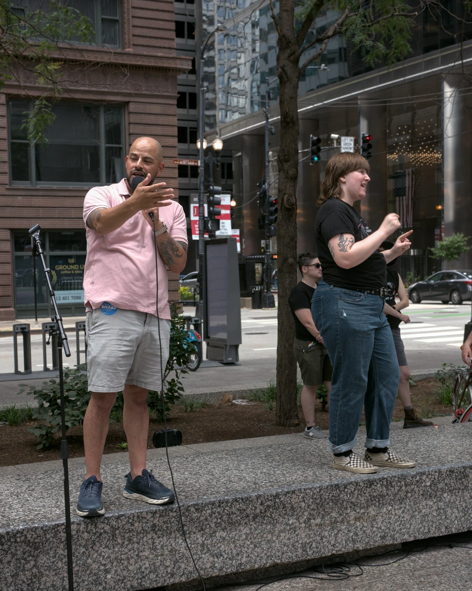   Alderman Andre Vasquez, a bald, tattooed, and bearded masc figure wearing a pink polo and shorts with black sneakers, stands next to Evryn Sevcech, who is wearing jeans, slides, and a black Trans Up Front shirt. Andre is addressing the crowd, and Evryn is translating his speech into ASL and is clearly having the time of their life doing so.  